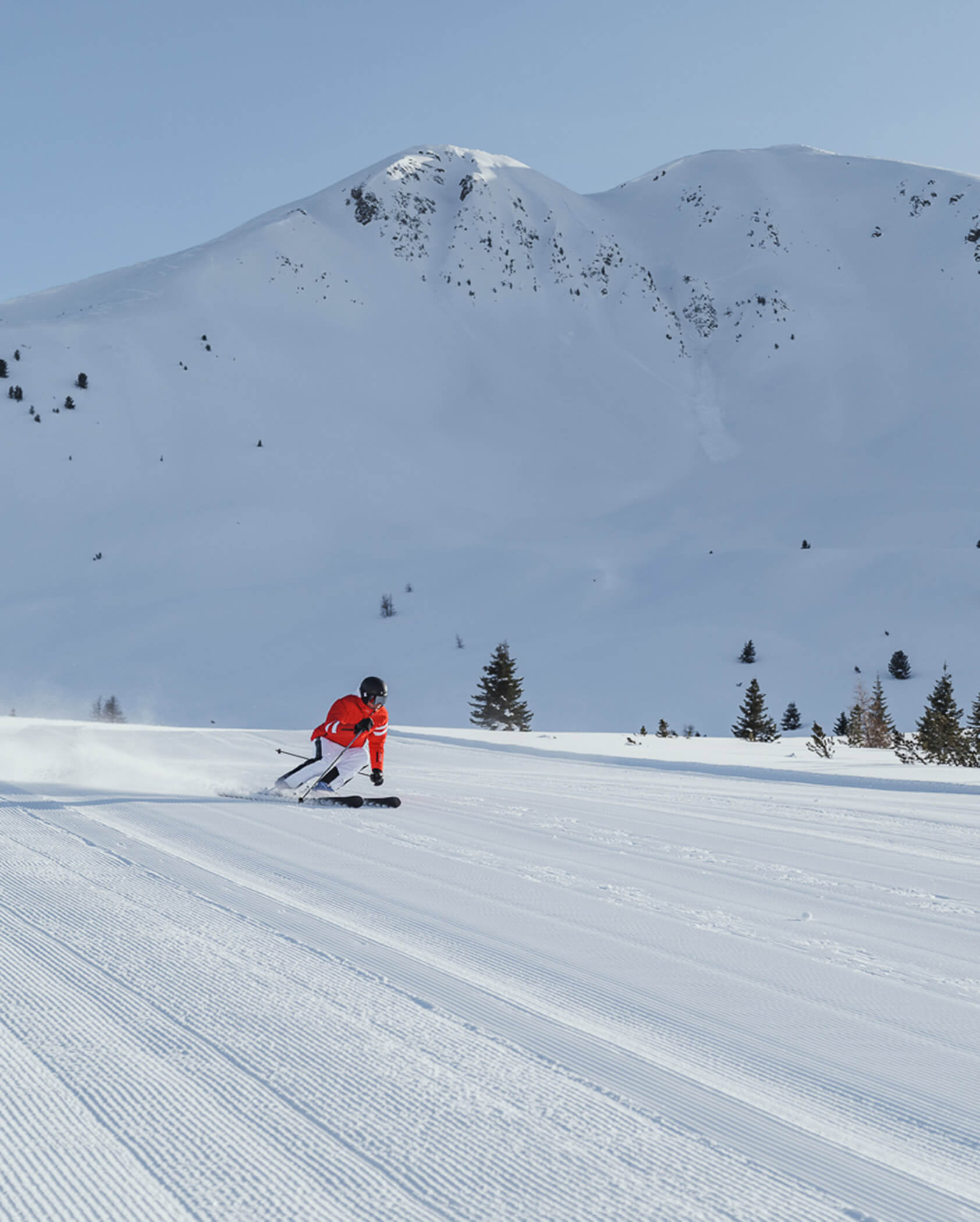 A skier in a red jacket skis down a groomed slope with snowy mountains in the background. - Hotel Tenz