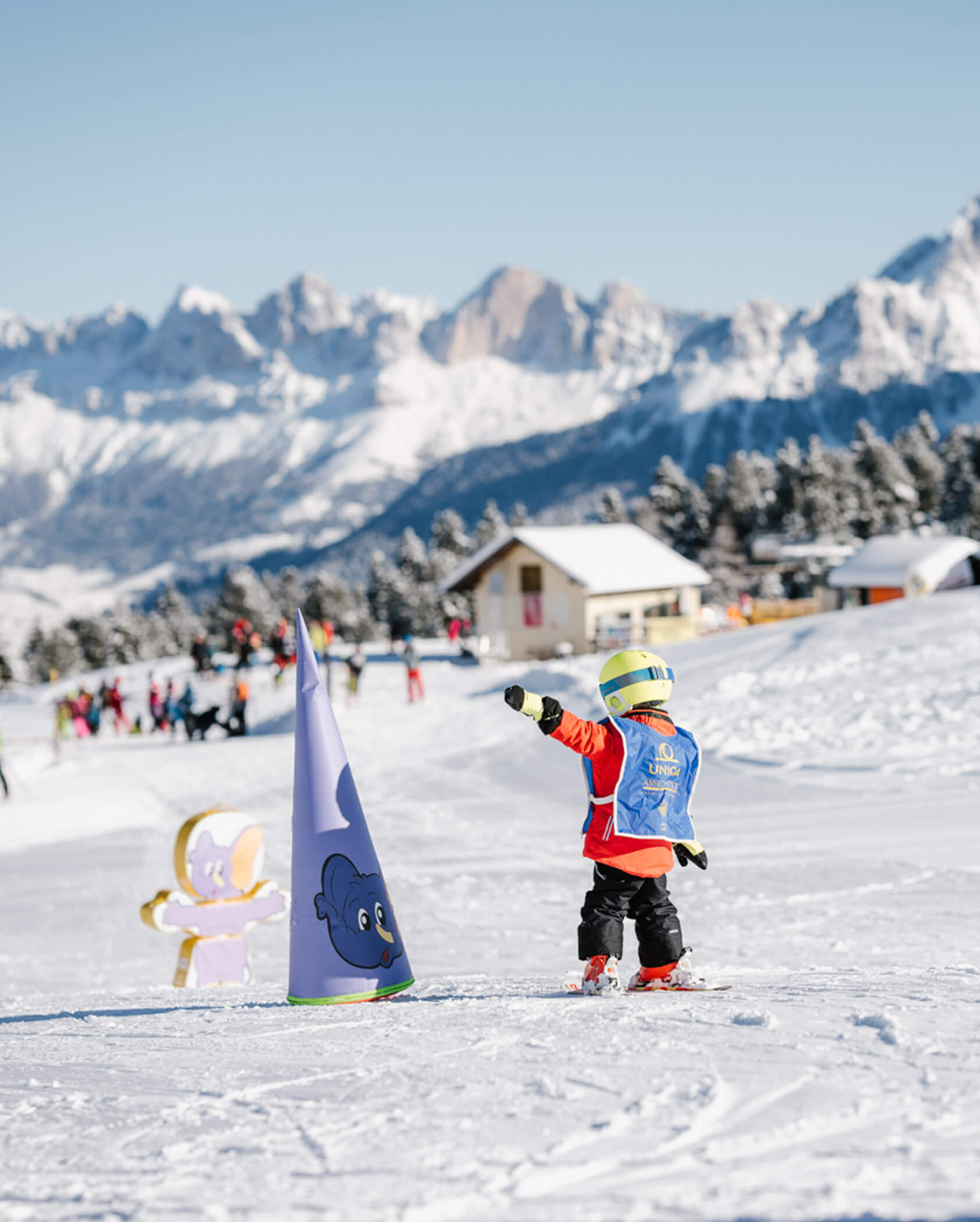 A child in ski gear skis on a snowy slope with mountains and a chalet in the background. - Hotel Tenz