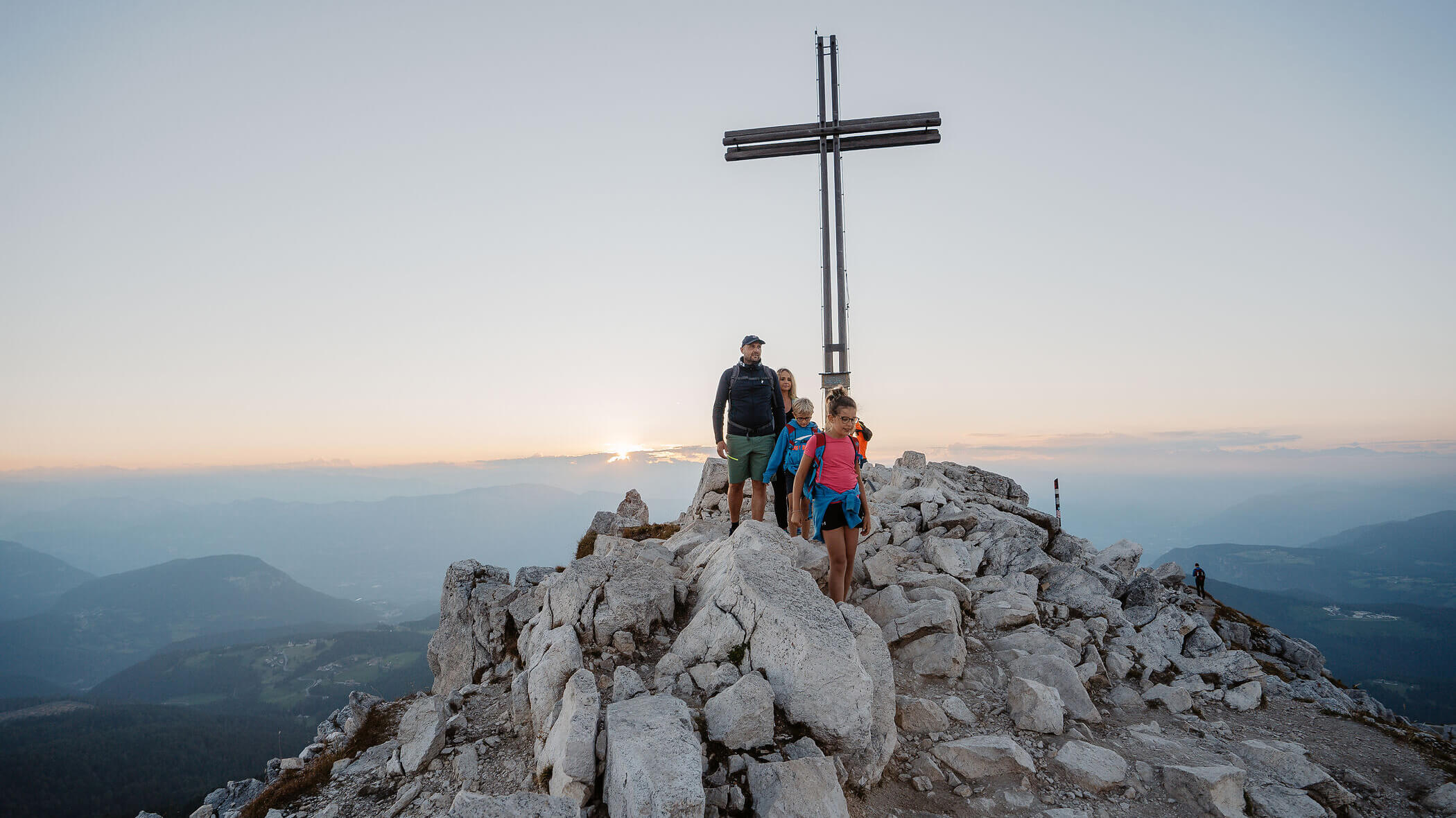 Four hikers stand on a rocky mountain peak beside a large cross at sunrise or sunset. - Hotel Tenz