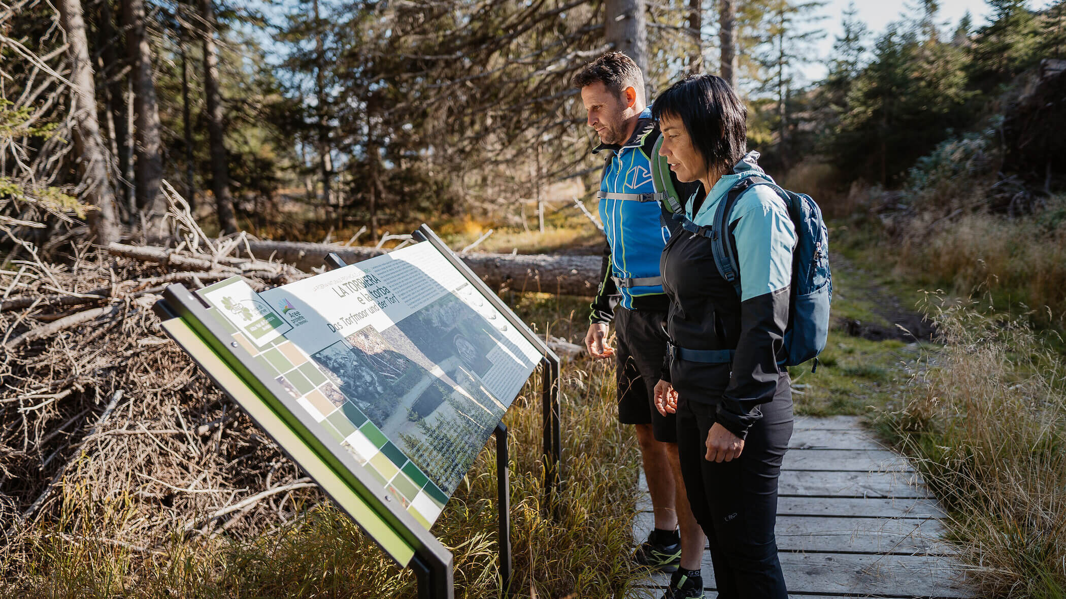 Two hikers read an informational sign on a forest trail surrounded by trees and greenery. - Hotel Tenz