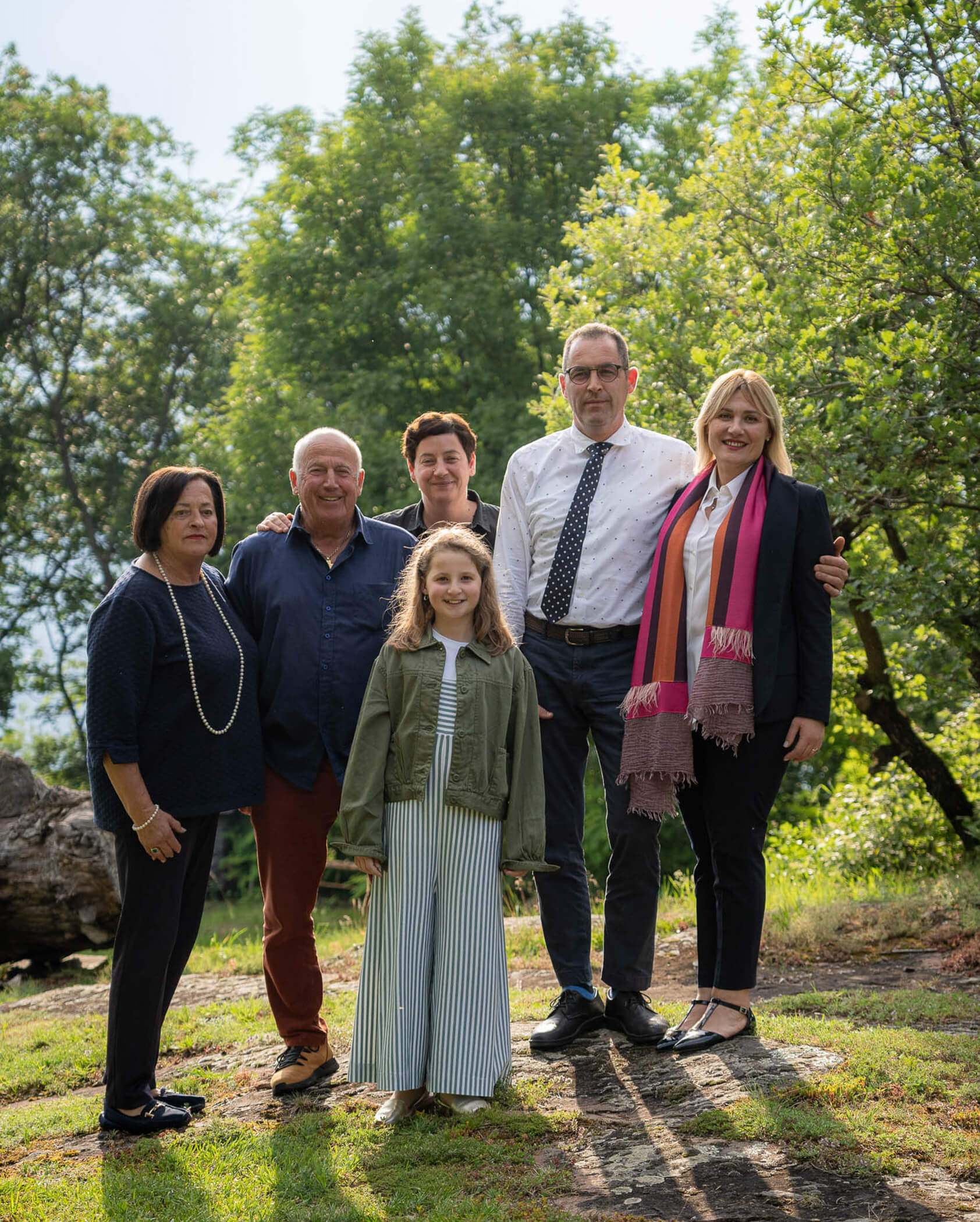 Five adults and one child pose outdoors on grass with trees in the background, all smiling at the camera. - Hotel Tenz