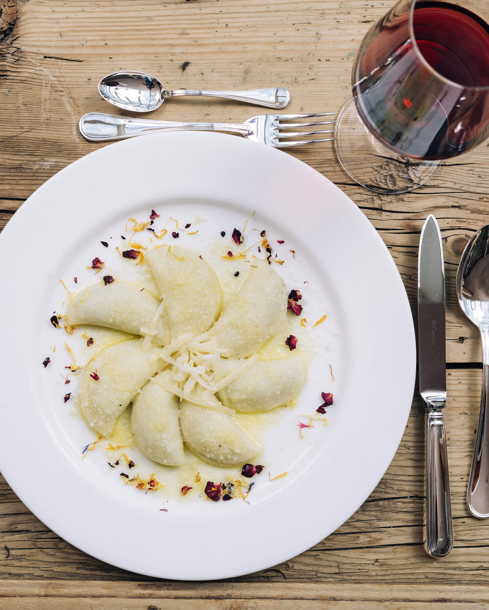 A plate of crescent-shaped ravioli with cheese and flower petals, beside cutlery and a glass of red wine. - Hotel Tenz