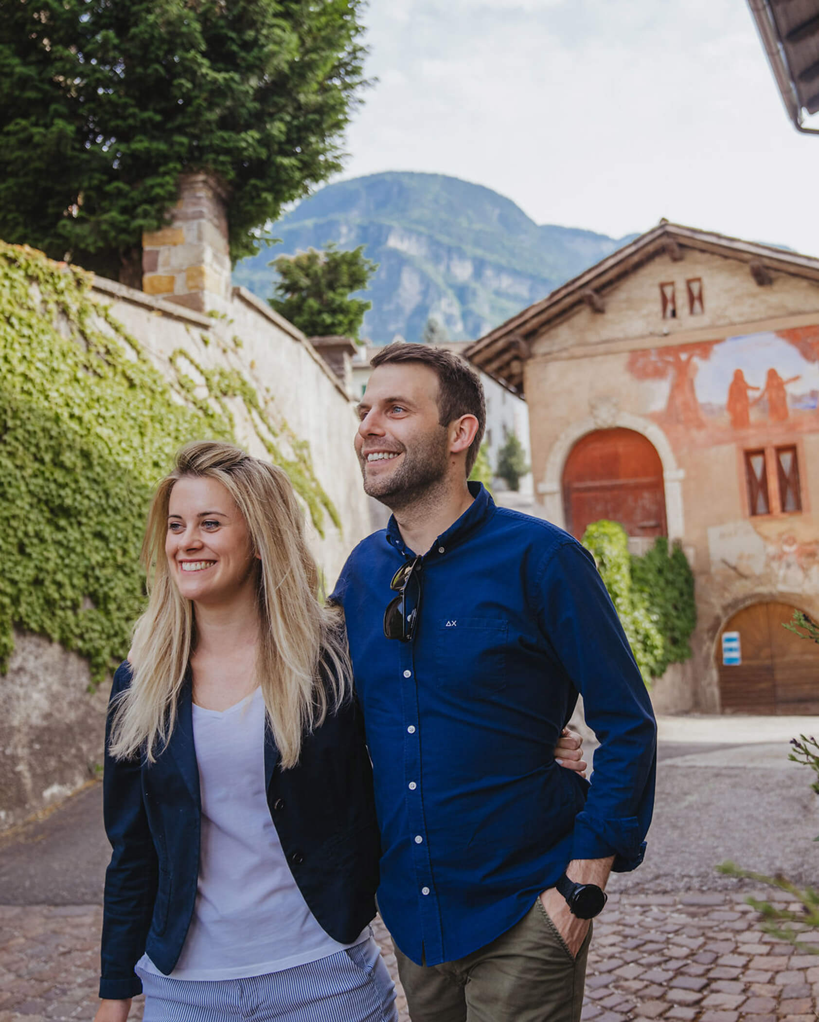 Smiling couple walking arm in arm on a cobblestone street with old buildings and mountains in the background. - Hotel Tenz