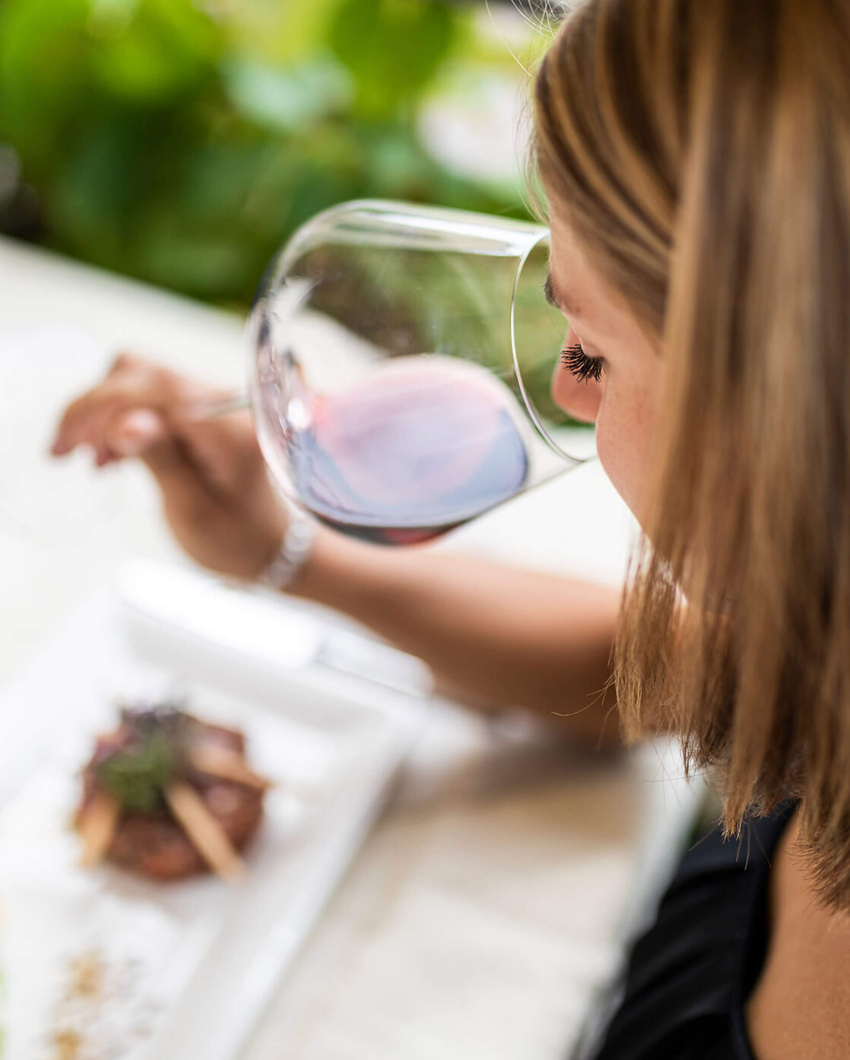 Woman with light brown hair smelling red wine in a glass at a table with a plated meal. - Hotel Tenz