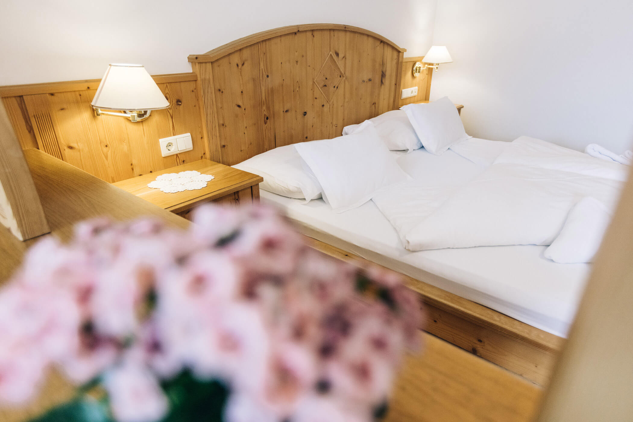 A cozy hotel room with a wooden bed, white bedding, bedside lamps, and pink flowers in the foreground. - Hotel Tenz