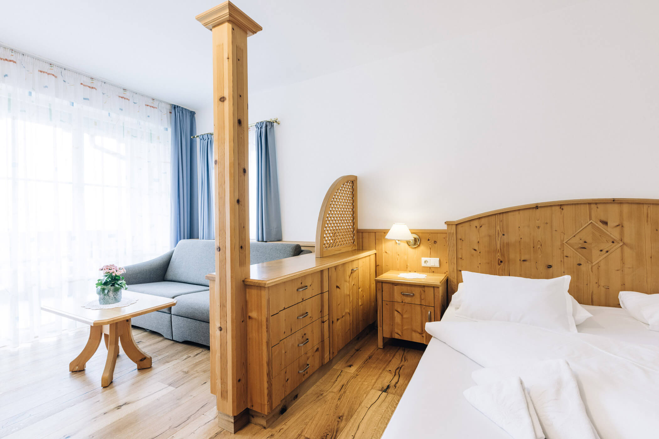 Bright hotel room with wooden furniture, a bed, sofa, side table, and soft natural light from large windows. - Hotel Tenz