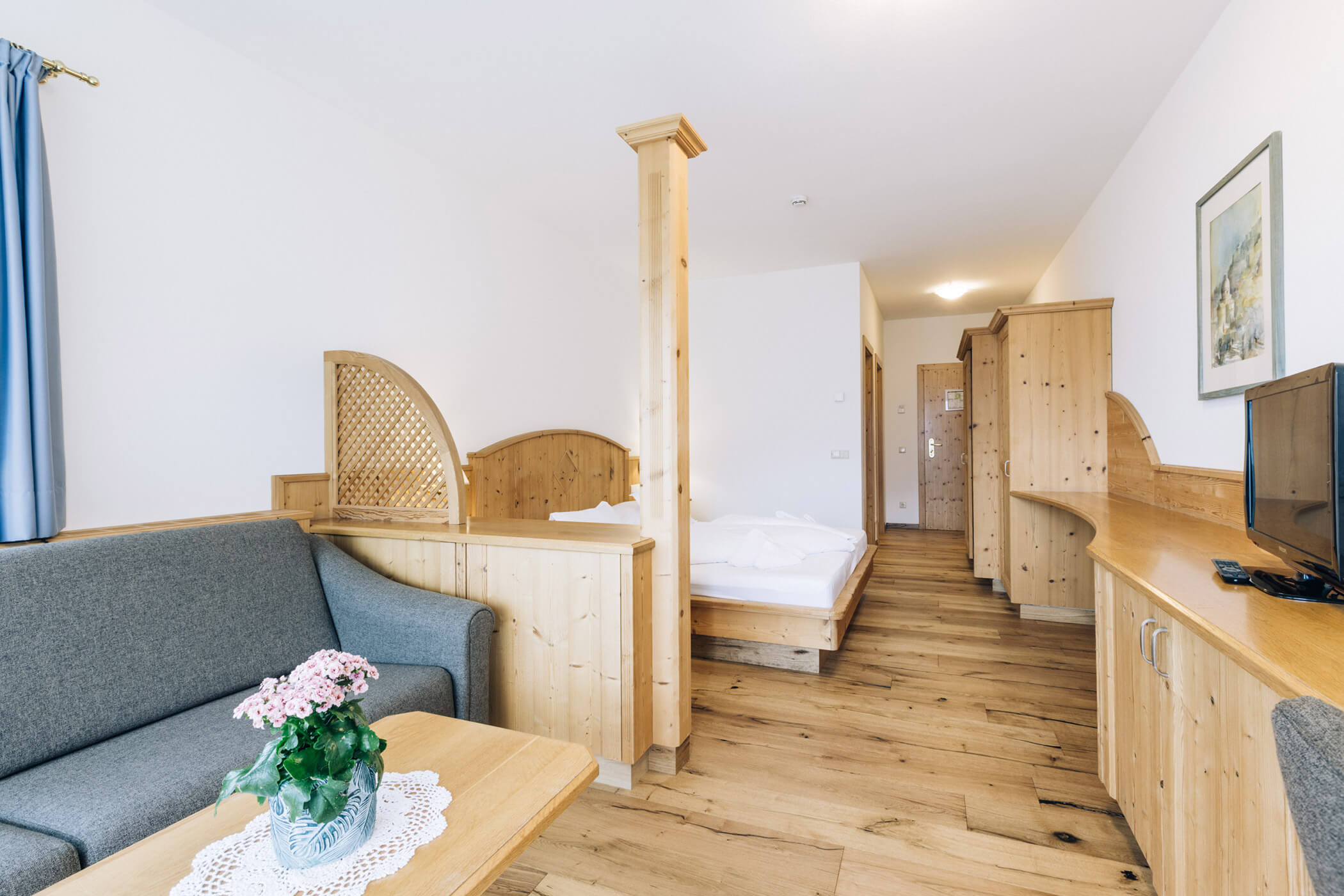 A bright hotel room with wooden furniture, a bed, sofa, TV, and a table with a pink flower vase. - Hotel Tenz