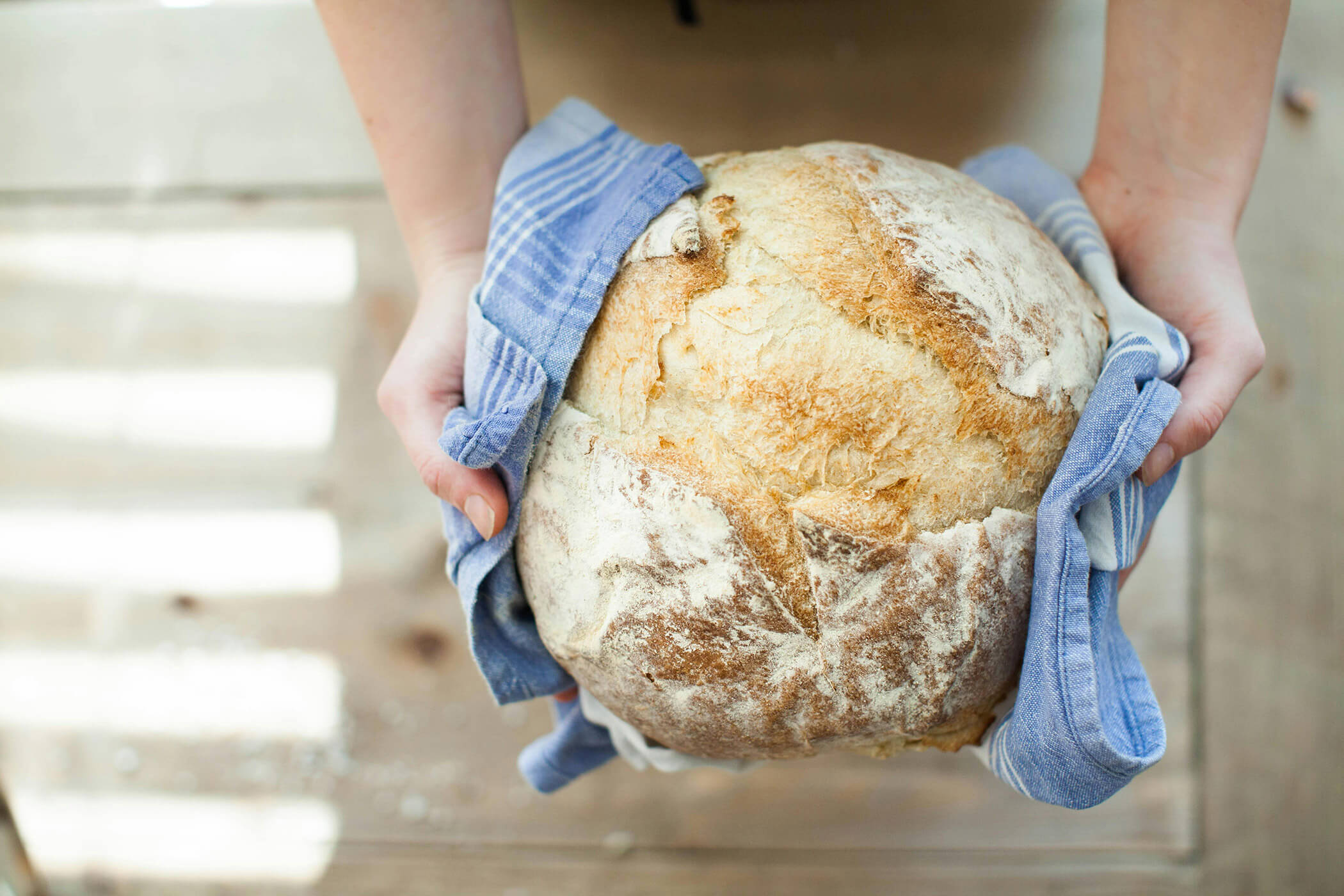 Hands holding a round loaf of crusty bread wrapped in a blue kitchen towel. - Hotel Tenz