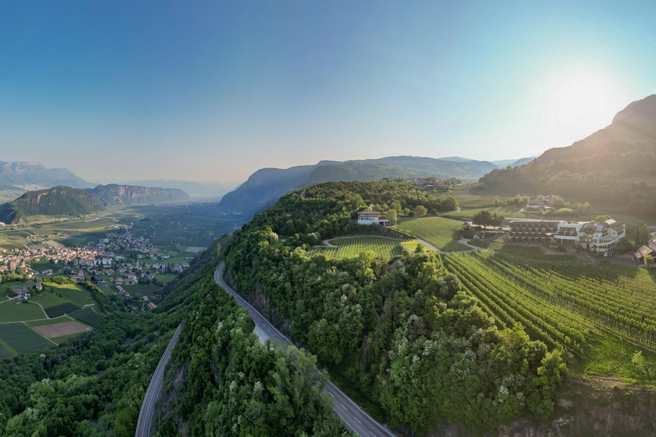 A winding road through green hills and vineyards with villages in a valley under a clear blue sky at sunset. - Hotel Tenz
