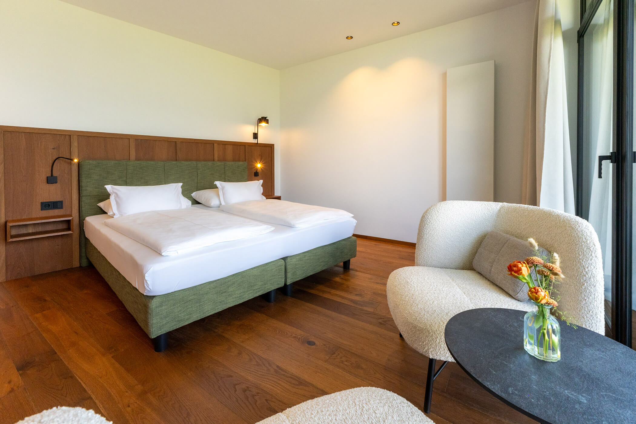 Modern hotel room with double beds, wooden floor, a cozy chair, and a small table with flowers in a vase. - Hotel Tenz