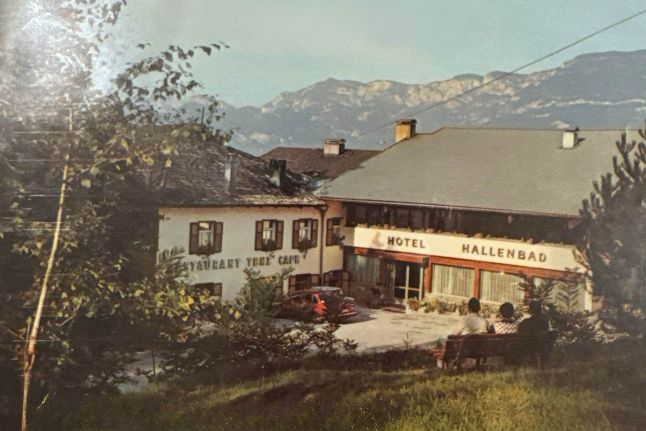 Zwei Personen sitzen auf einer Bank mit Blick auf ein Hotel und ein Restaurant, im Hintergrund sind die Berge zu sehen. - Hotel Tenz