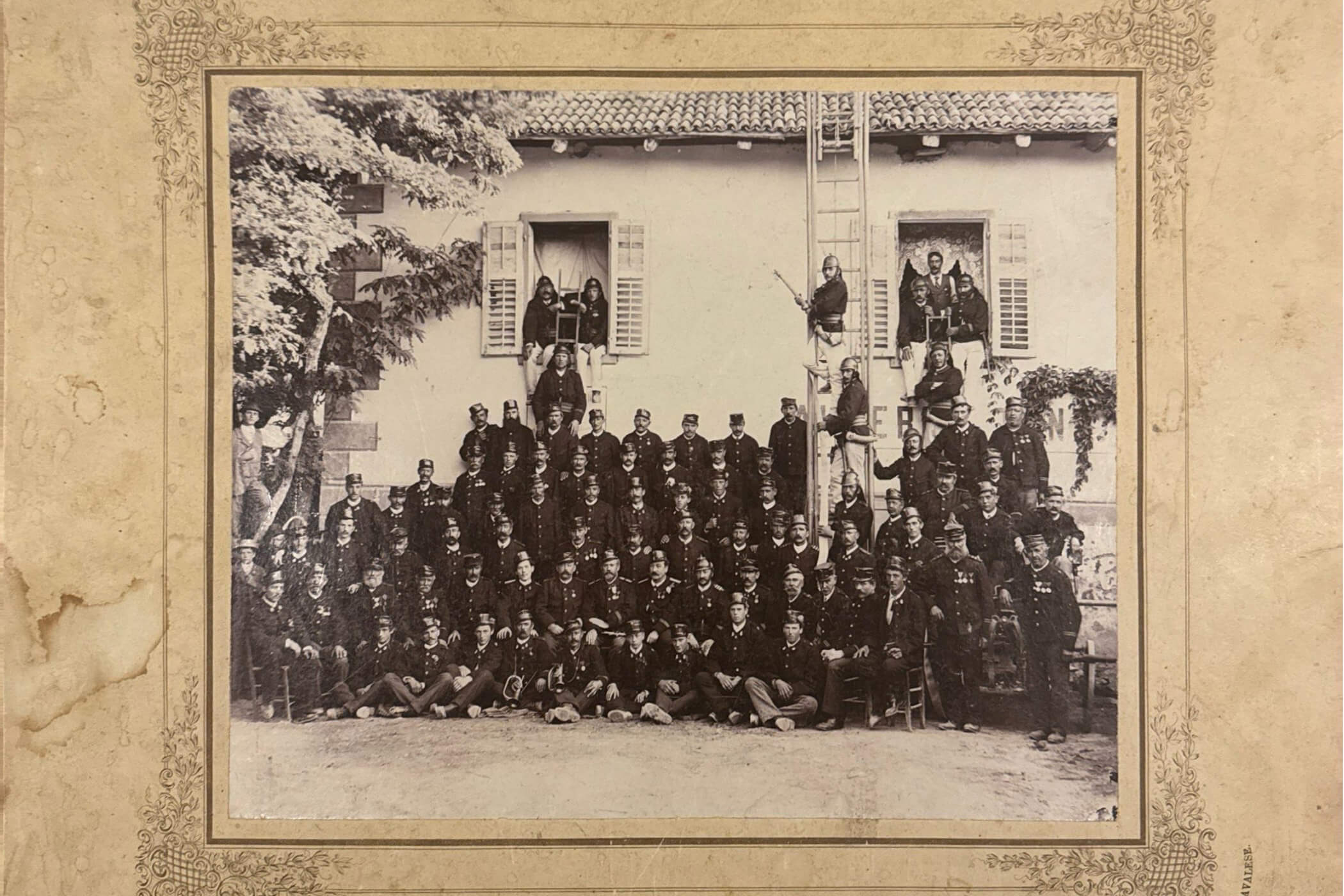 Eine große Gruppe von Uniformierten posiert vor einem Gebäude; einige sitzen in Fenstern, andere stehen auf einer Leiter. - Hotel Tenz