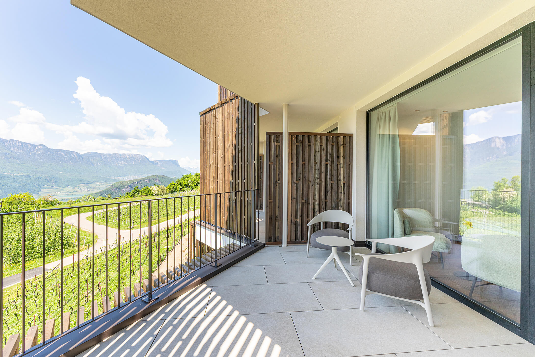Moderner Balkon mit zwei Stühlen und einem Tisch, mit Blick auf Weinberge und Berge unter einem sonnigen Himmel. - Hotel Tenz