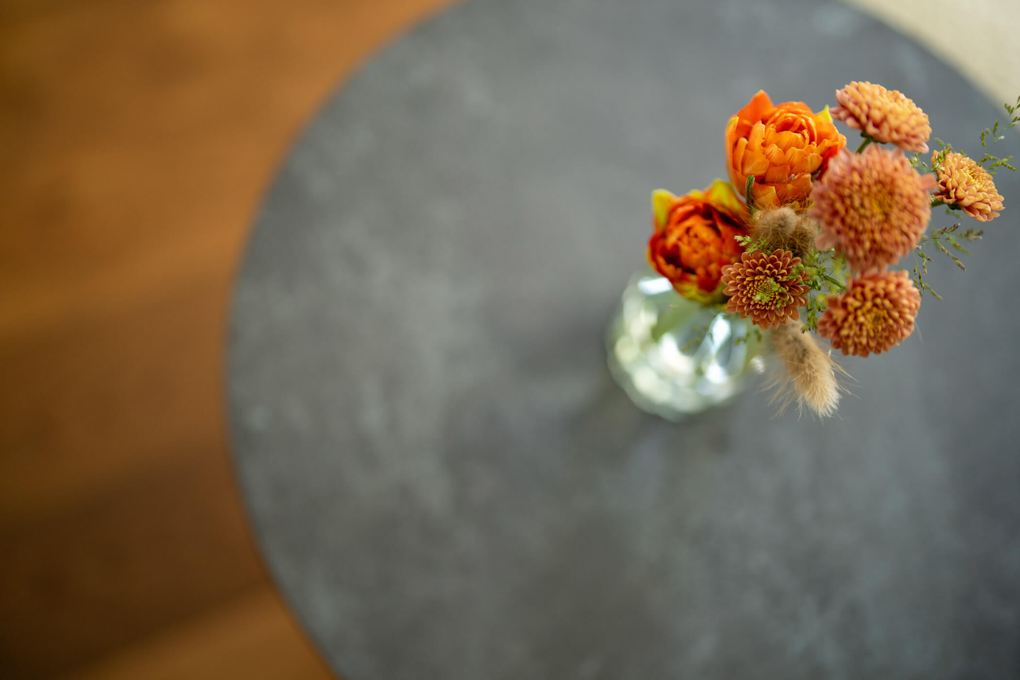 Orangefarbene Blumen in einer Glasvase auf einem runden, dunkelgrauen Tisch, von oben gesehen. - Hotel Tenz