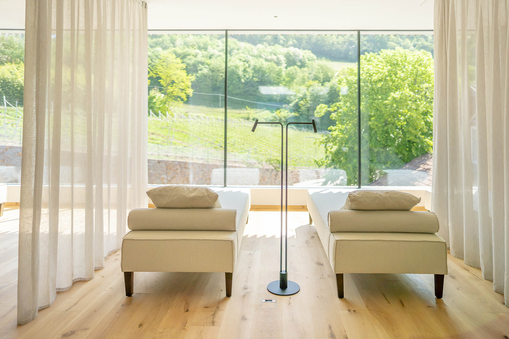 Zwei beigefarbene Liegen stehen vor großen Glasfenstern mit Blick auf die grünen Hügel und Bäume. - Hotel Tenz