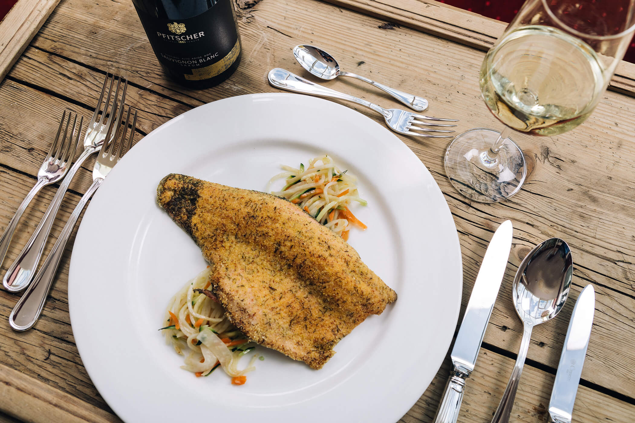 A breaded fish fillet on vegetables, served with white wine and cutlery on a rustic wooden table. - Hotel Tenz