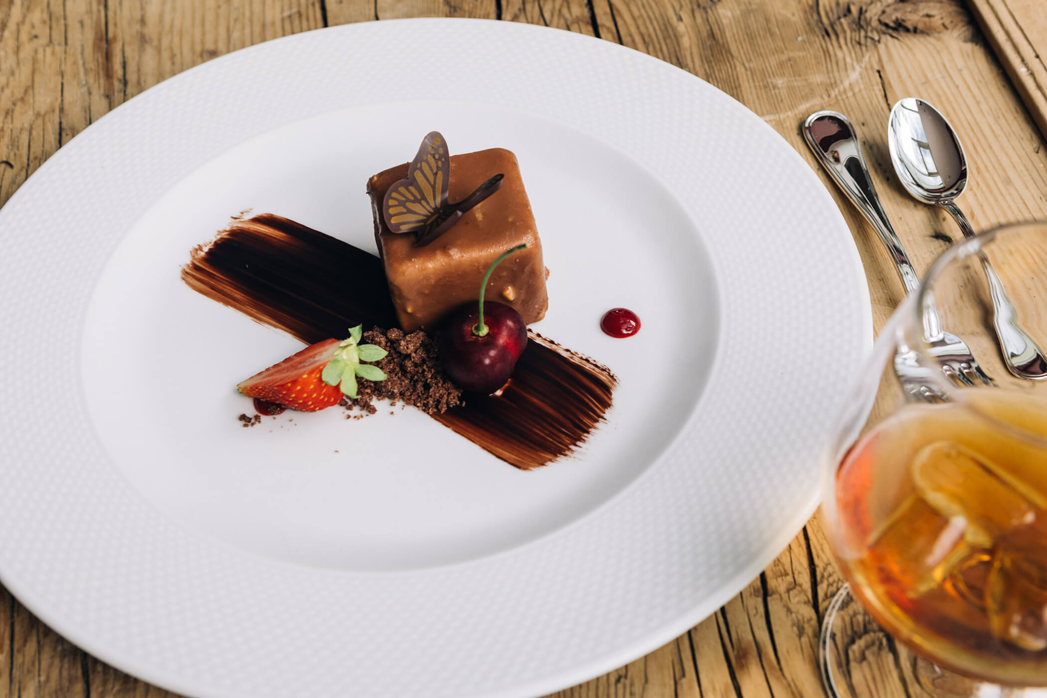 A plated chocolate dessert with a cherry, strawberry, and decorative chocolate butterfly on a wooden table. - Hotel Tenz