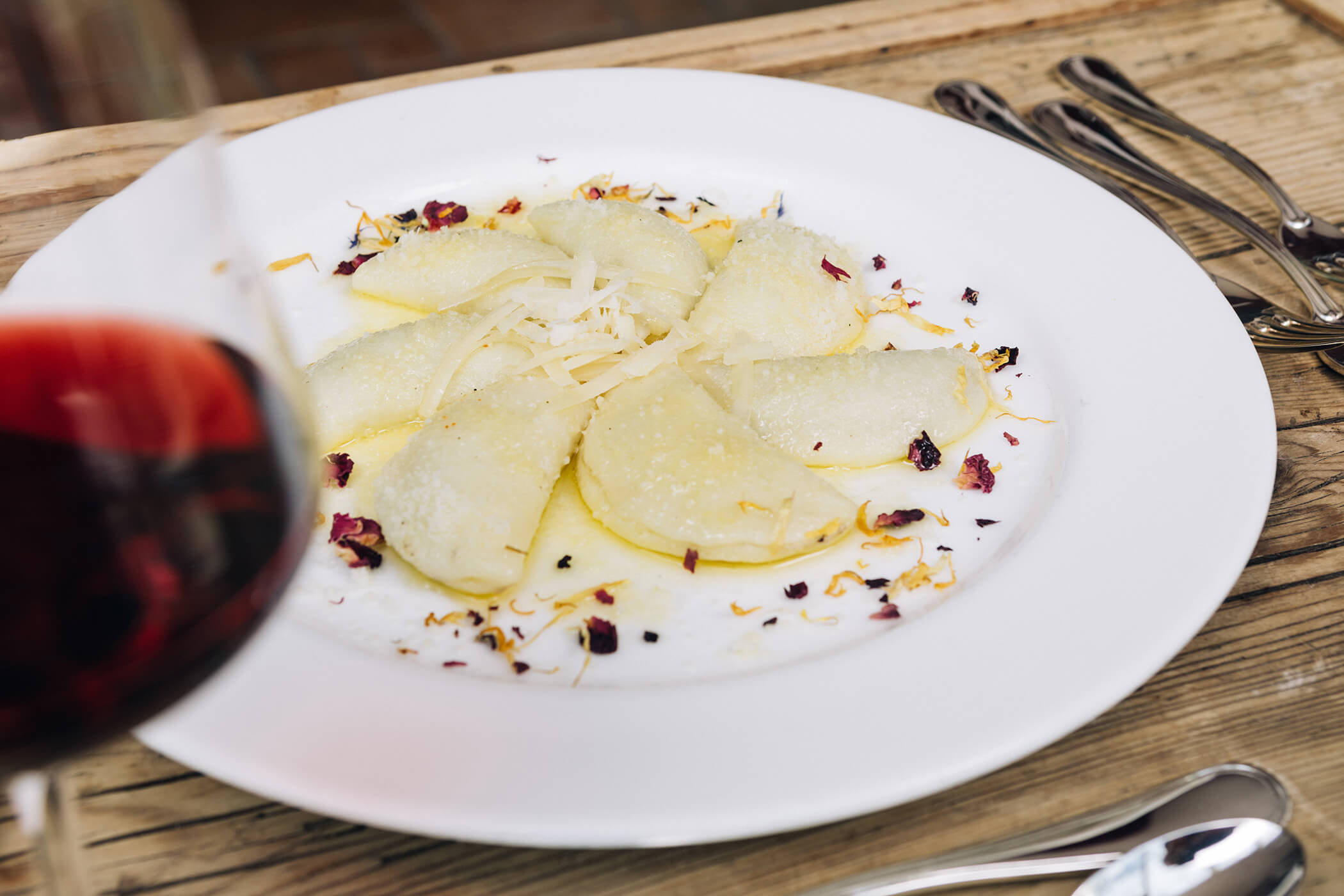 A white plate of ravioli with cheese and herbs, next to a glass of red wine on a wooden table. - Hotel Tenz