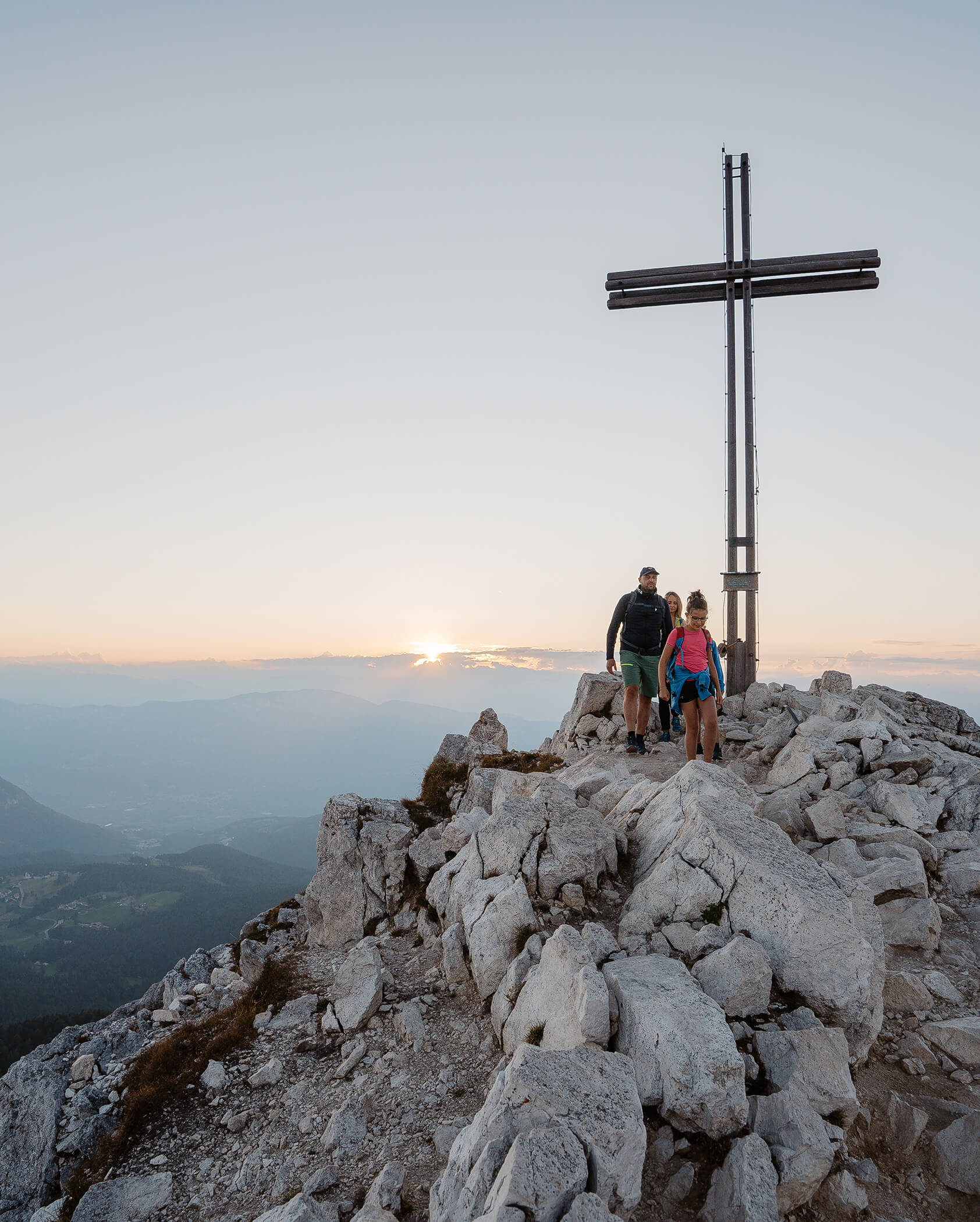 Zwei Wanderer stehen bei Sonnenaufgang oder Sonnenuntergang an einem großen Kreuz auf einem felsigen Berggipfel und überblicken ein Tal. - Hotel Tenz
