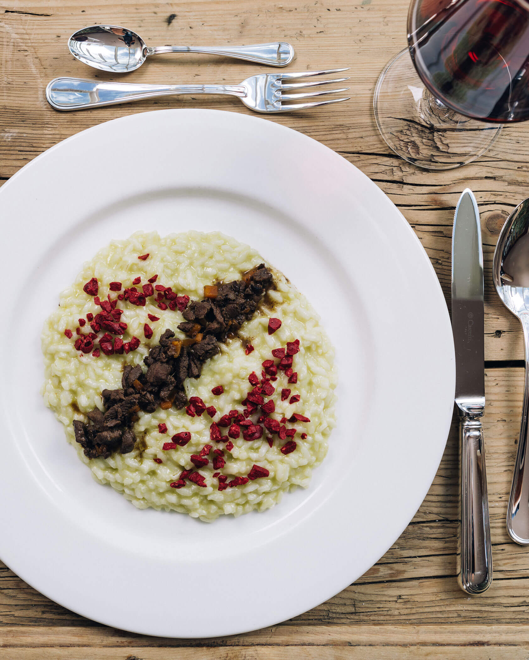 Ein Teller cremiges Risotto mit gewürfelter roter Beilage und einem Streifen Pilze, daneben ein Glas Rotwein. - Hotel Tenz