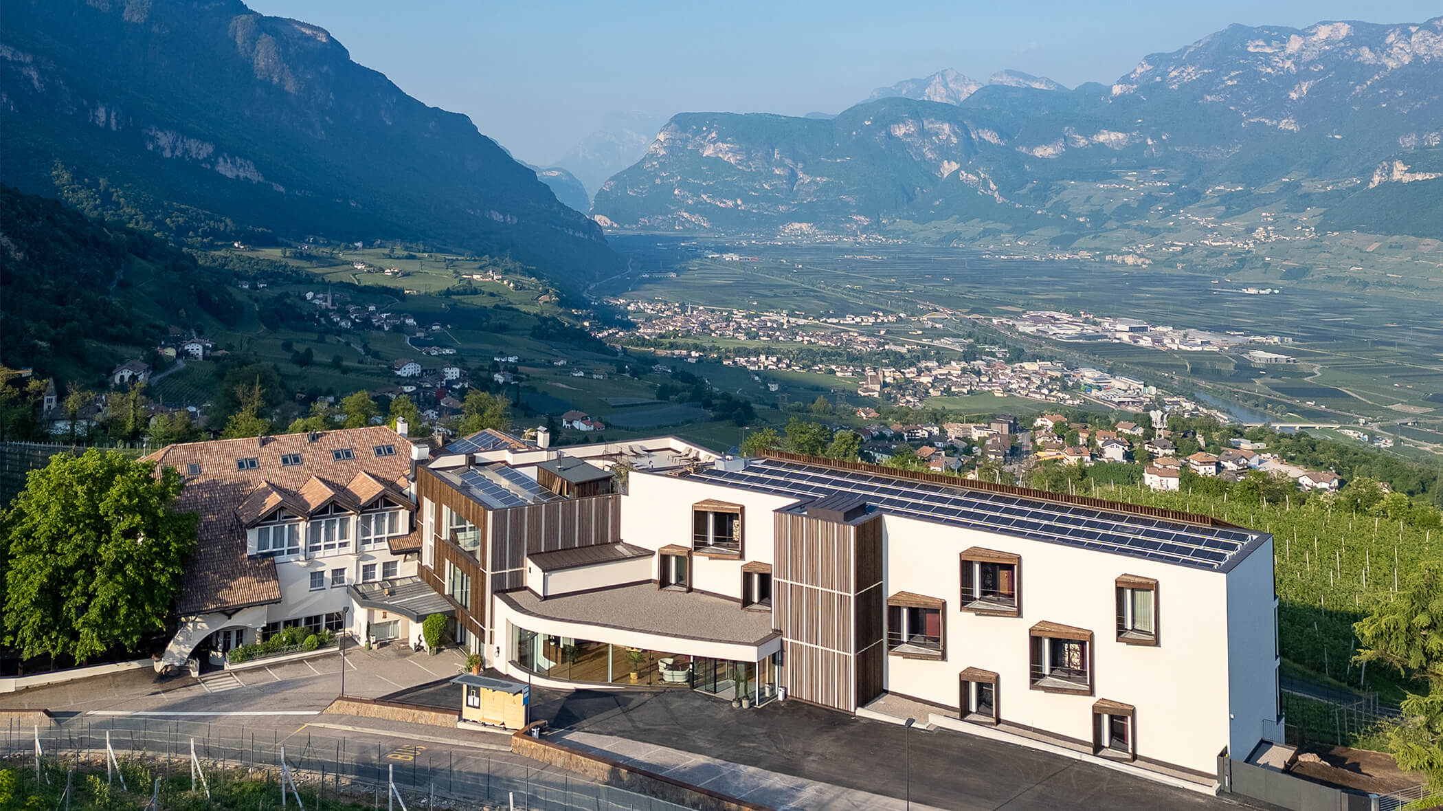 Modern building with solar panels overlooking a valley, vineyards, and mountains under a clear sky. - Hotel Tenz