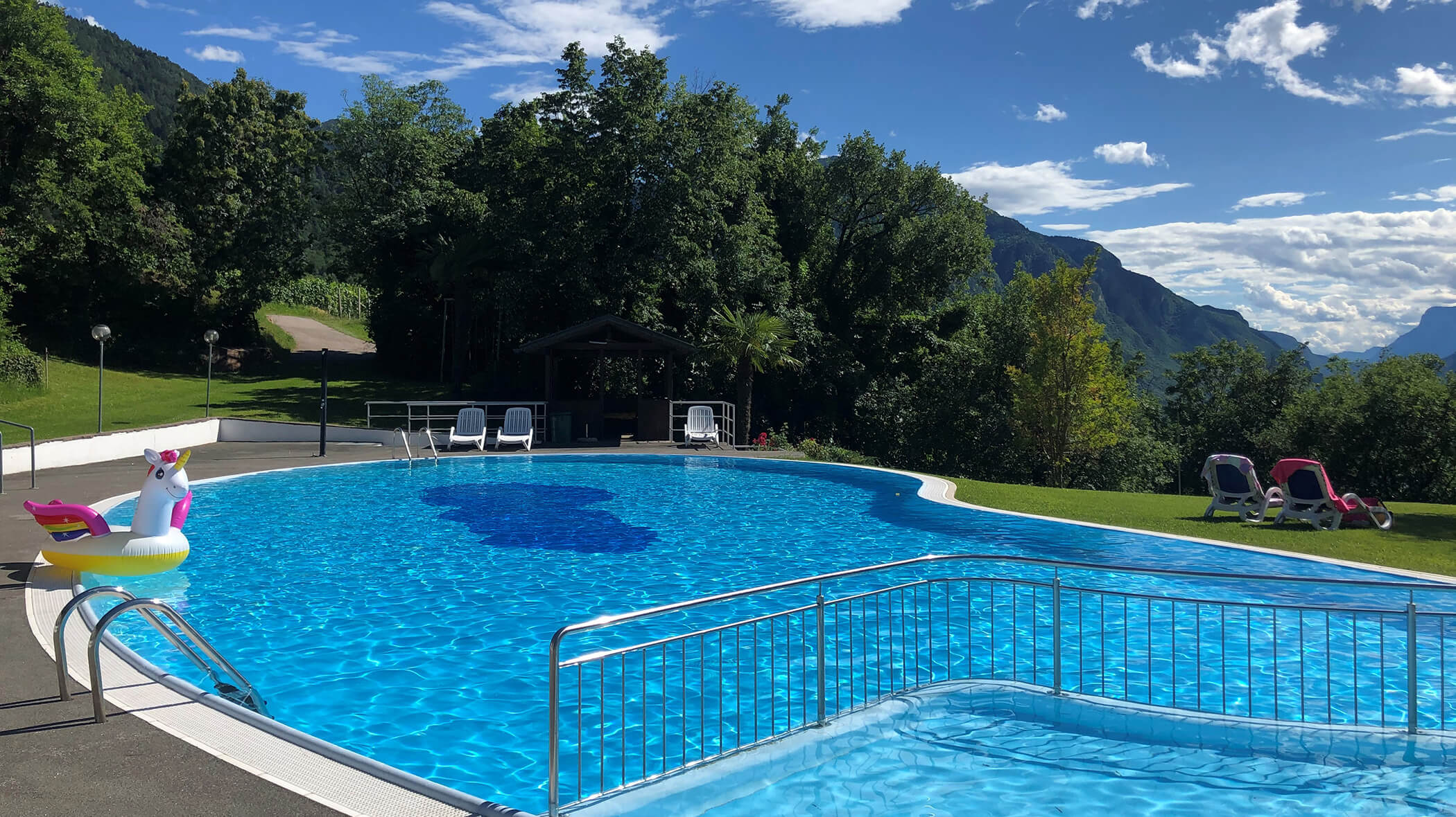 Outdoor swimming pool with clear blue water, surrounded by greenery and mountains, with pool chairs and a unicorn float. - Hotel Tenz