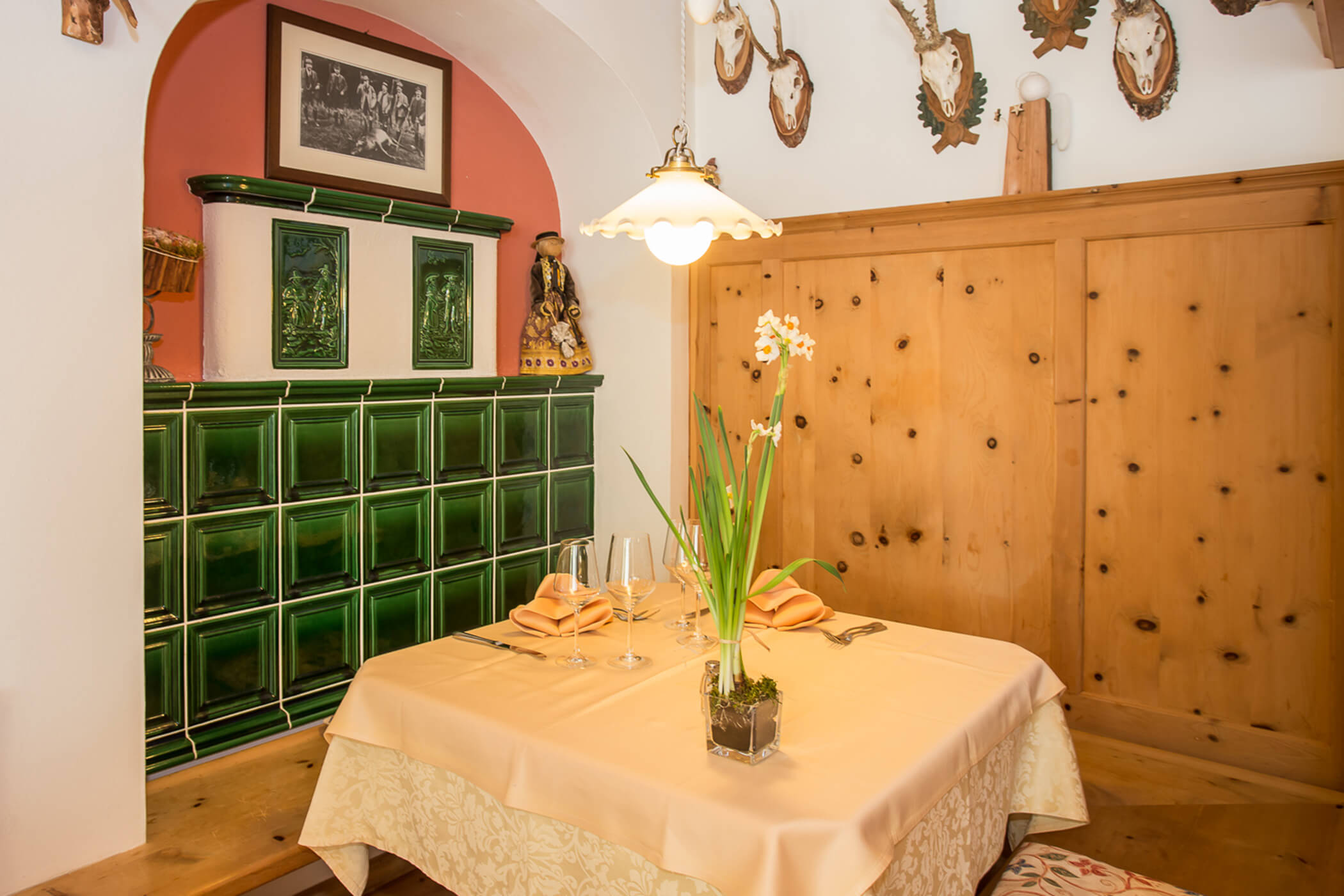 Cozy dining nook with a set table, green tile stove, wood paneling, and mounted antlers on the wall. - Hotel Tenz