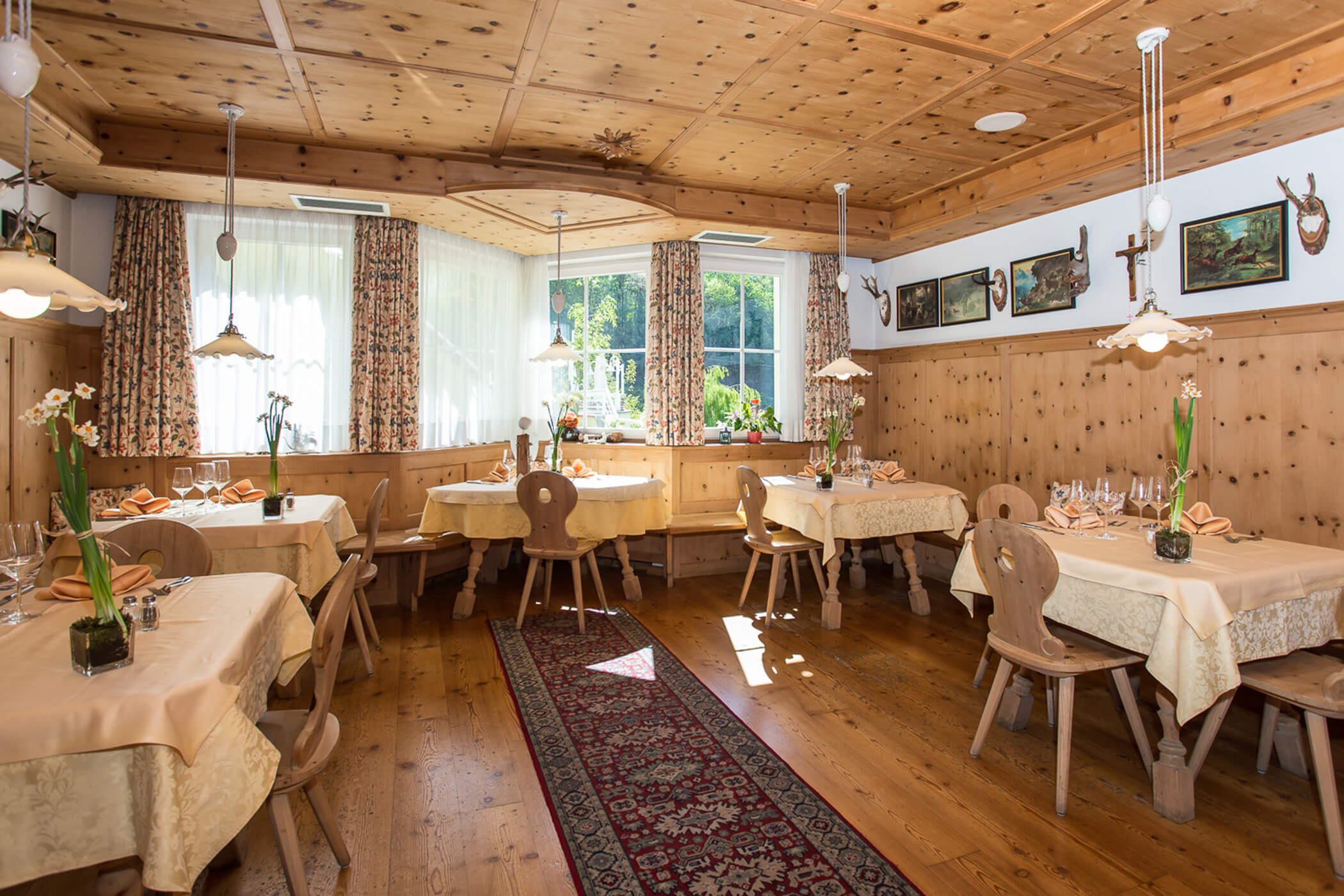 Cozy, sunlit dining room with wooden furniture, cream tablecloths, and deer antler wall decor. - Hotel Tenz
