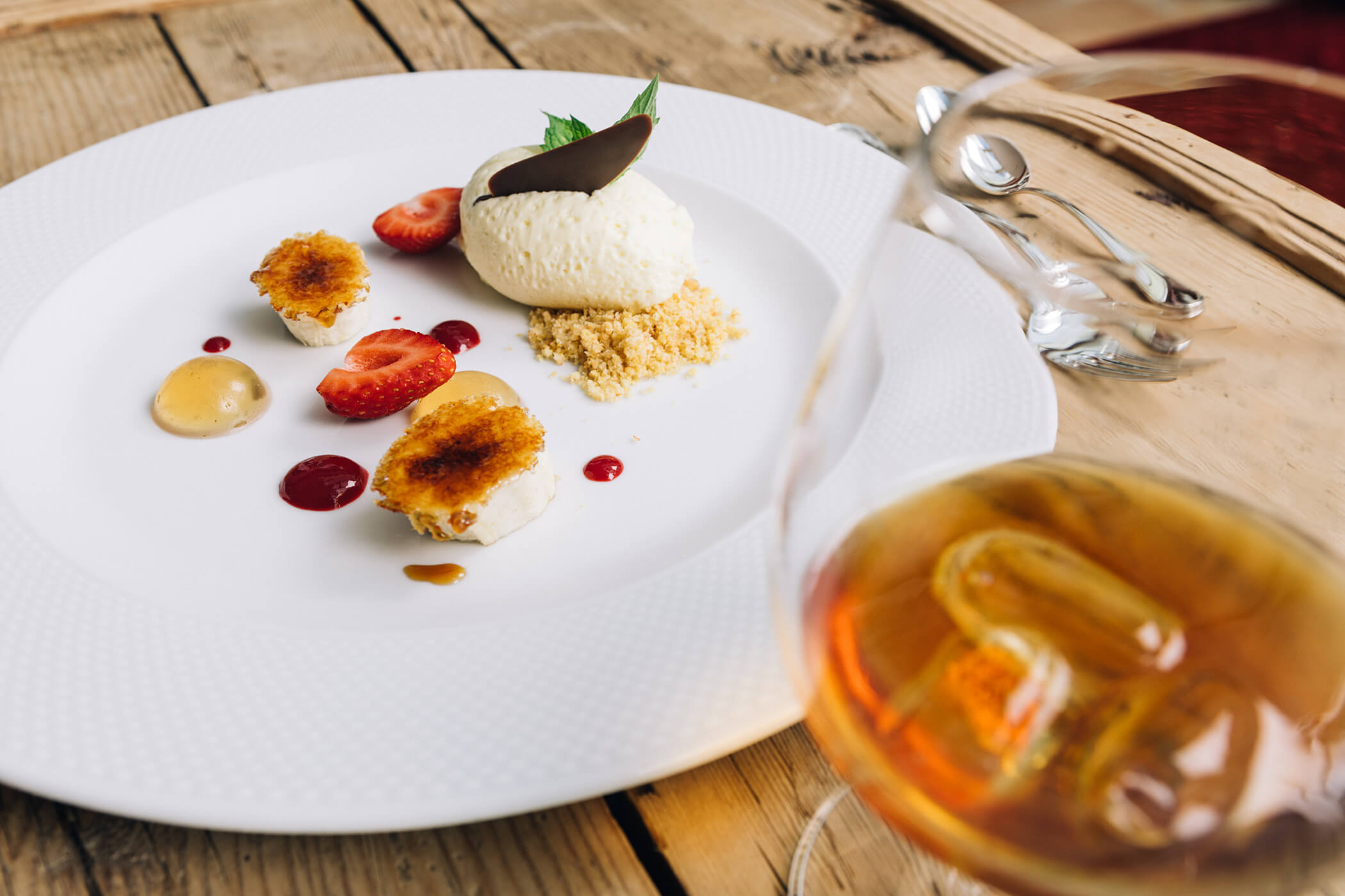 A gourmet dessert with strawberries, cream, and crumble on a white plate, with a glass of iced drink nearby. - Hotel Tenz