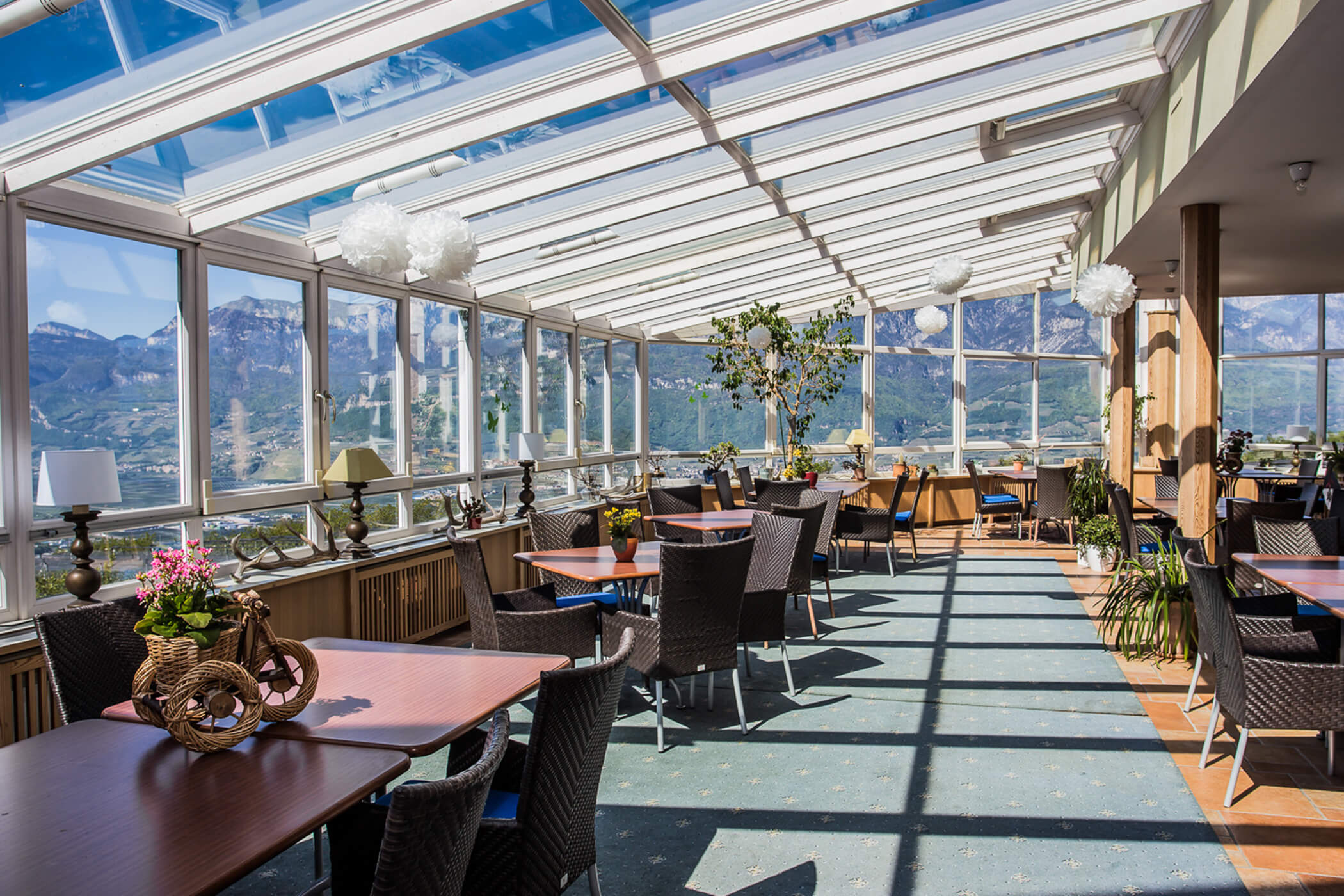 Sunlit conservatory restaurant with wicker chairs, potted plants, and mountain views through large windows. - Hotel Tenz