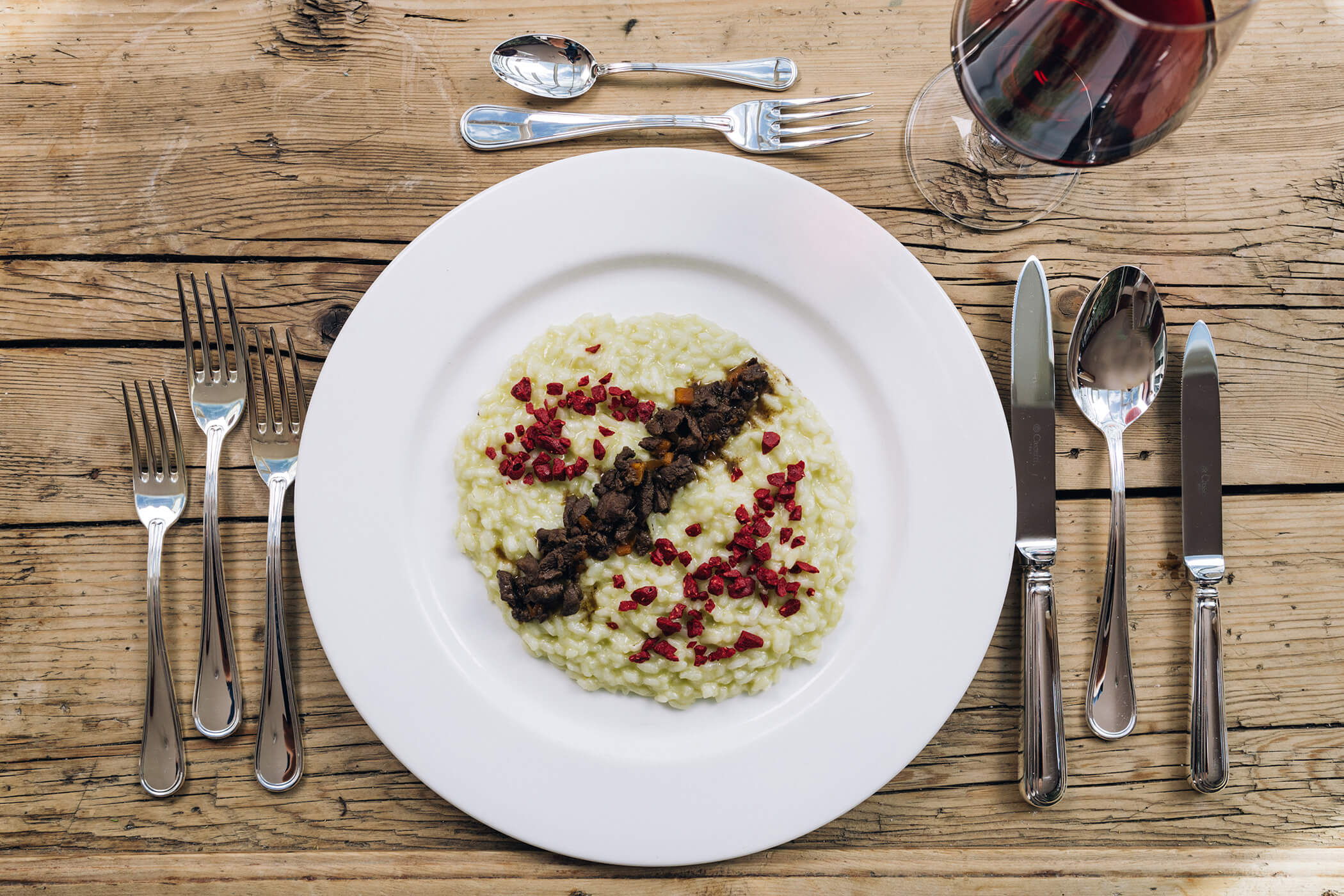 A plate of creamy risotto with garnishes, set on a rustic wooden table with cutlery and a glass of red wine. - Hotel Tenz