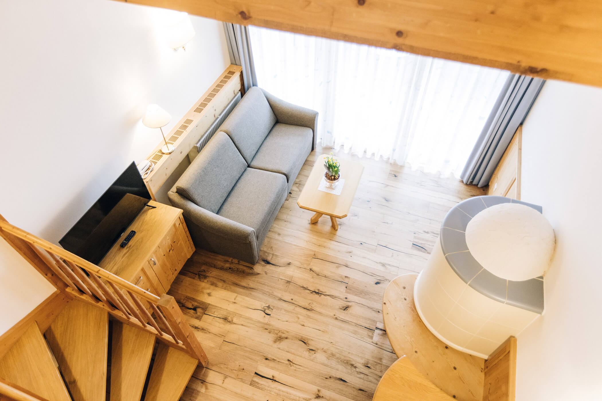Cozy living room with a gray sofa, wooden floors, small table, TV, and staircase, viewed from above. - Hotel Tenz