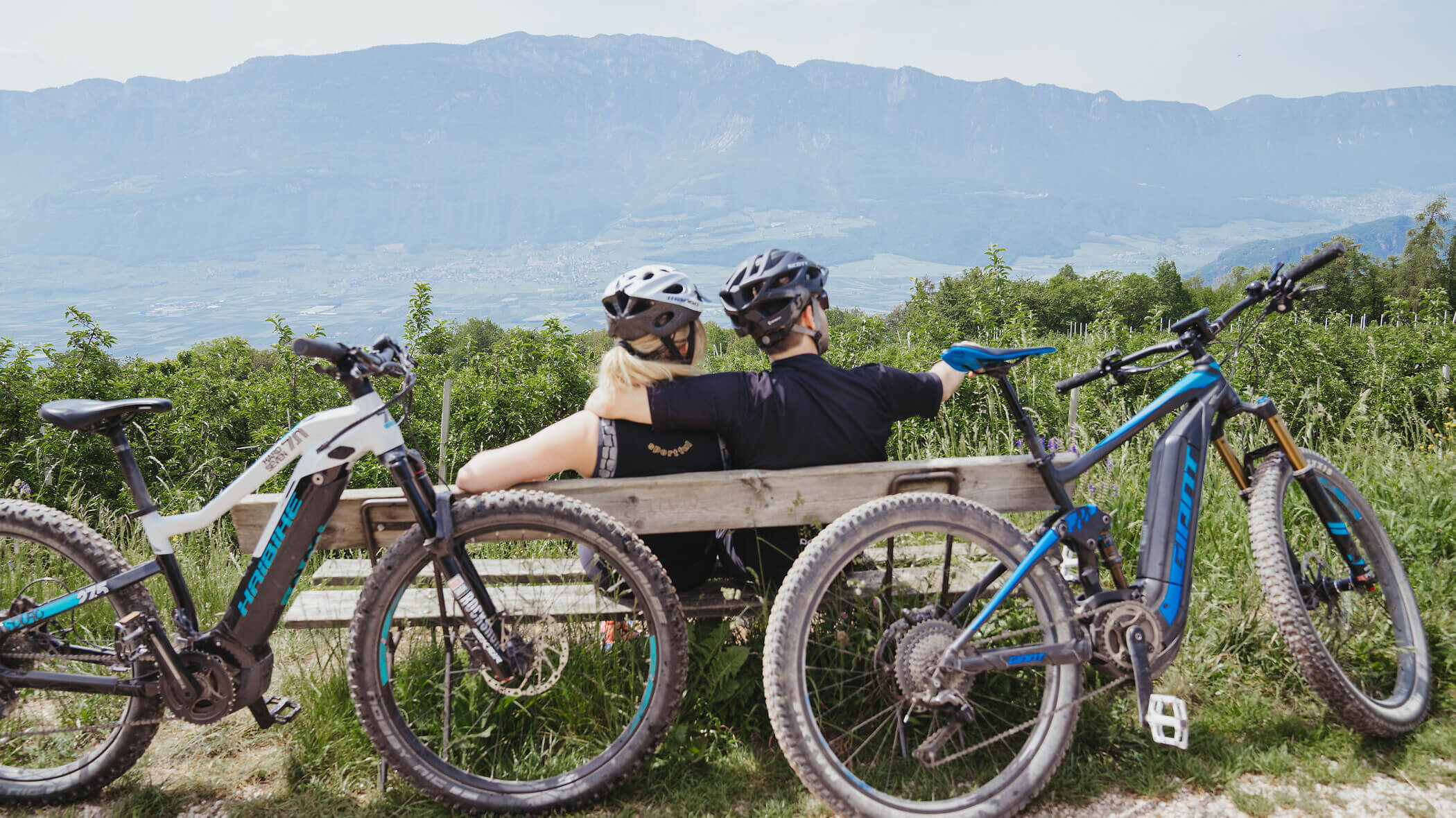 Two cyclists sit on a bench, backs to the camera, with bikes parked nearby and mountains in the background. - Hotel Tenz