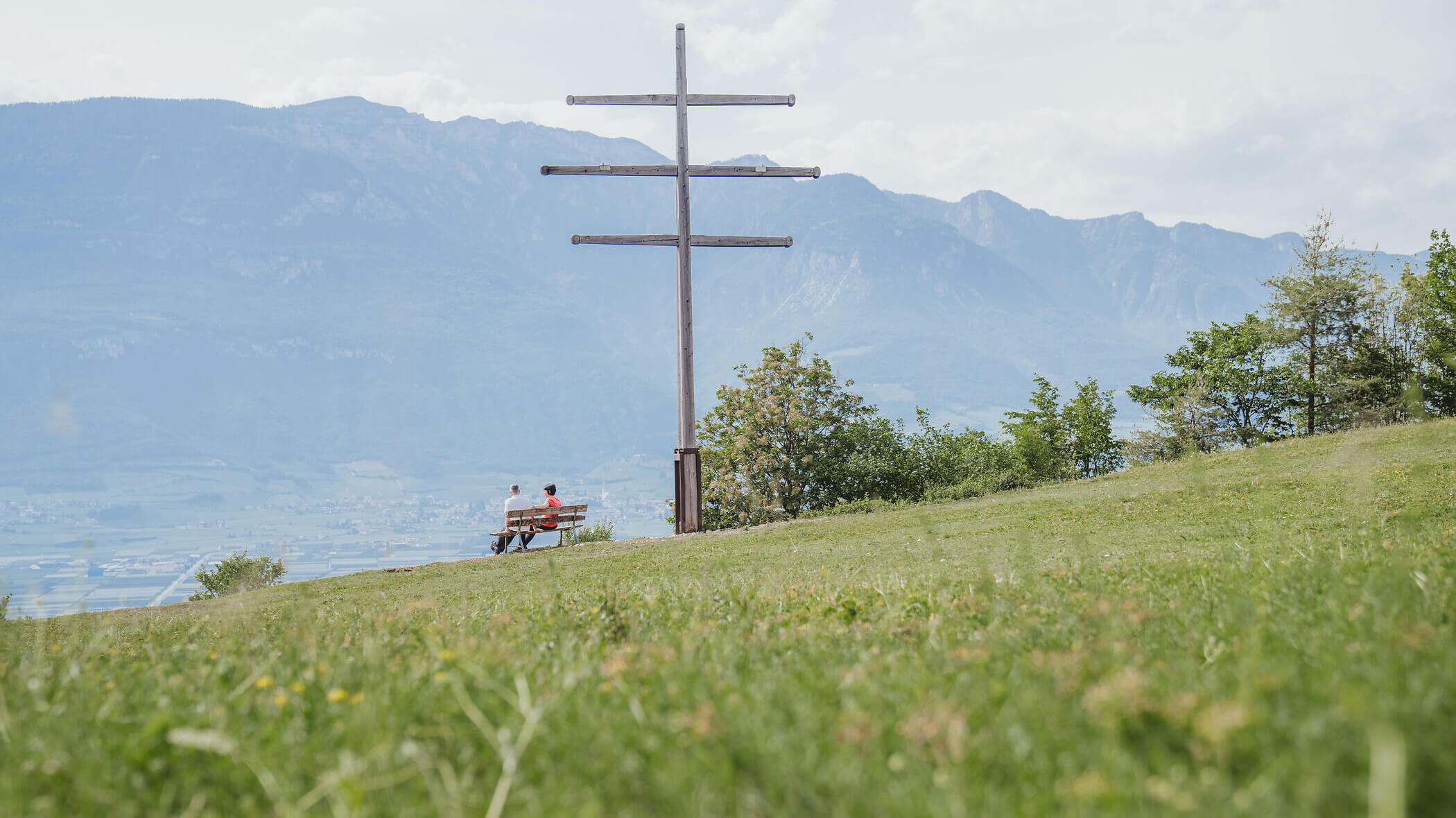 Two people sit on a bench near a large wooden cross with mountains in the background on a grassy hill. - Hotel Tenz