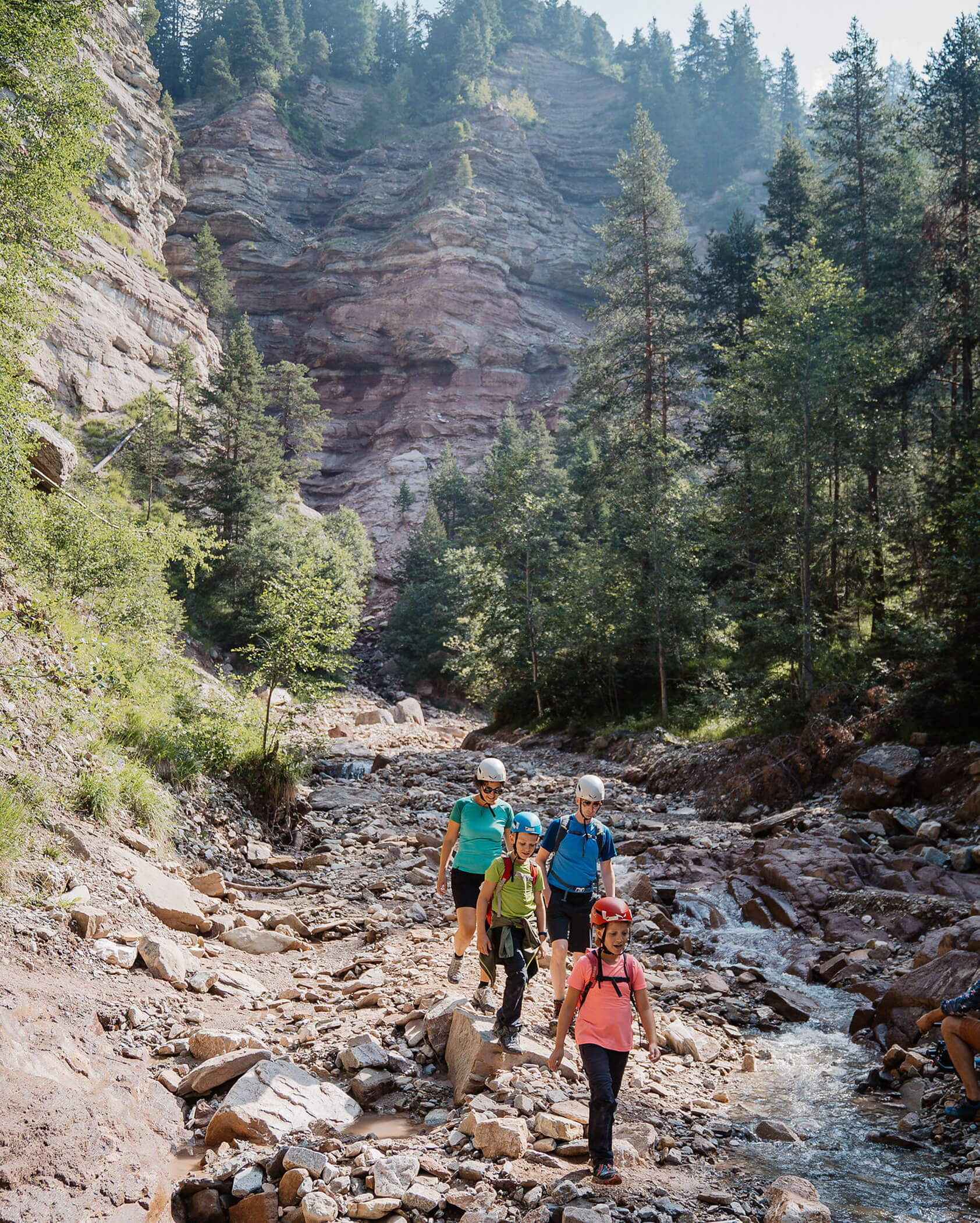 Four people hike along a rocky creek in a forested canyon with tall cliffs and trees in the background. - Hotel Tenz