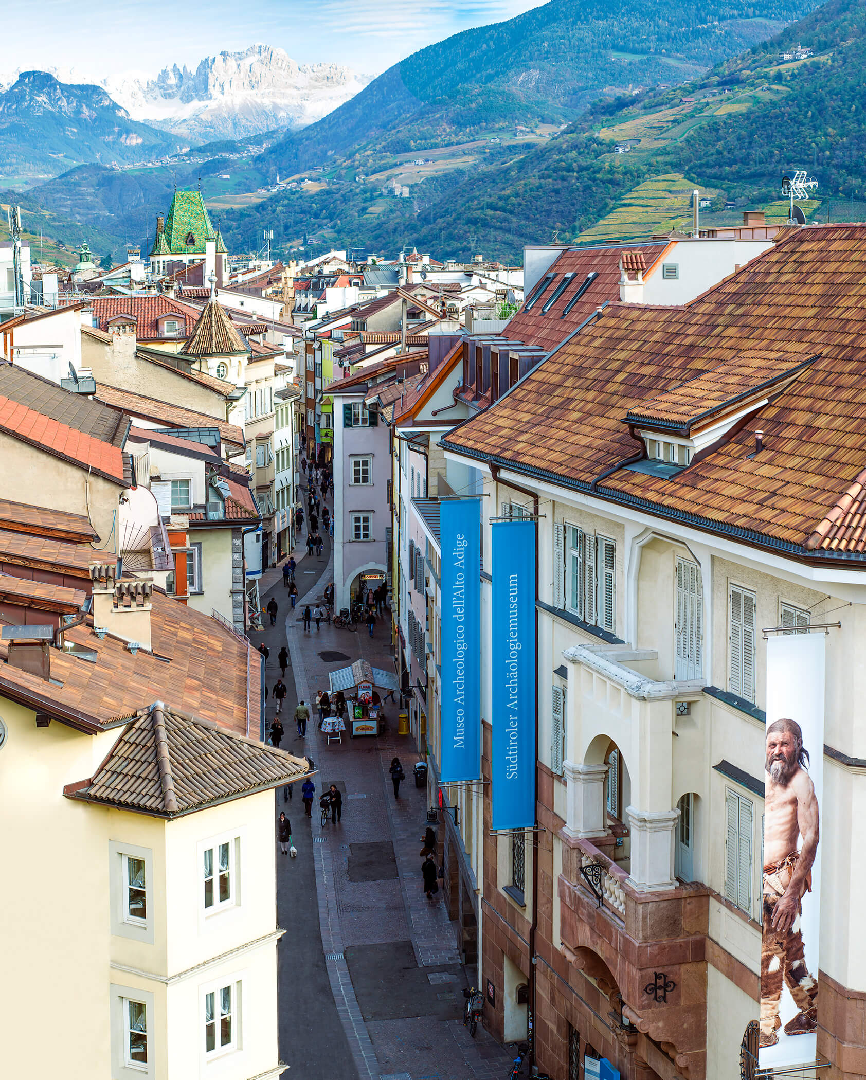 A scenic view of Bolzano’s old town with mountains in the background and banners on buildings. - Hotel Tenz