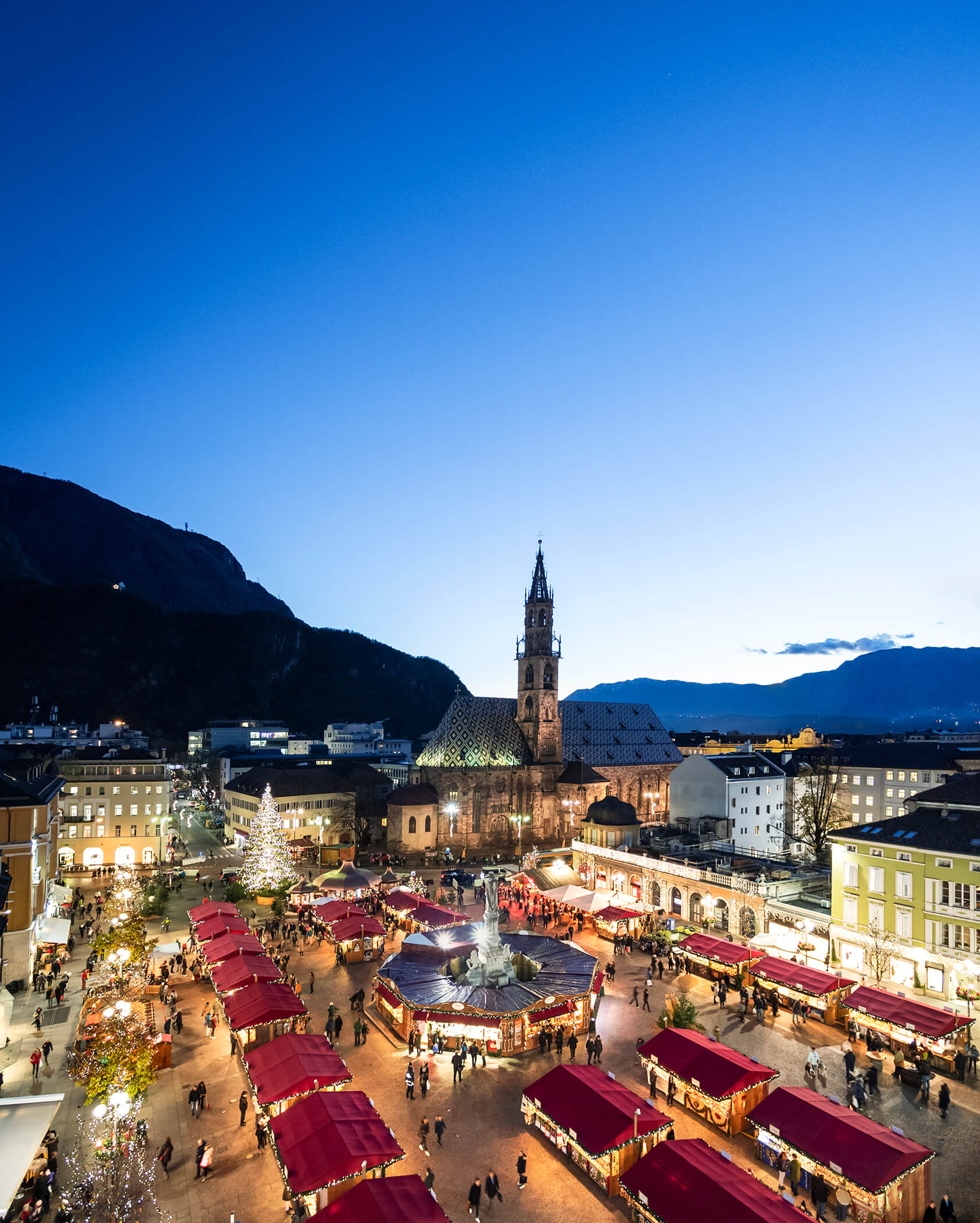 A festive Christmas market with red stalls and lights in a town square at dusk, mountains in the background. - Hotel Tenz
