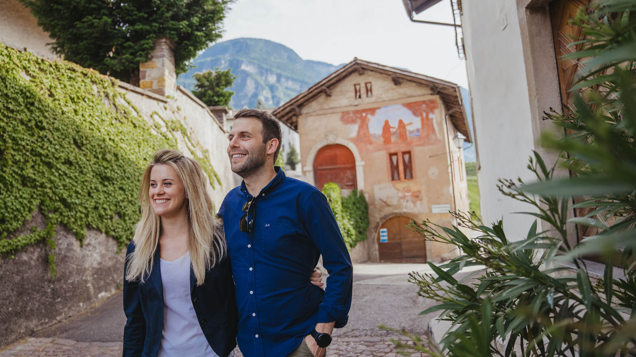 A smiling couple walks arm in arm down a charming, sunny street with historic buildings and greenery. - Hotel Tenz