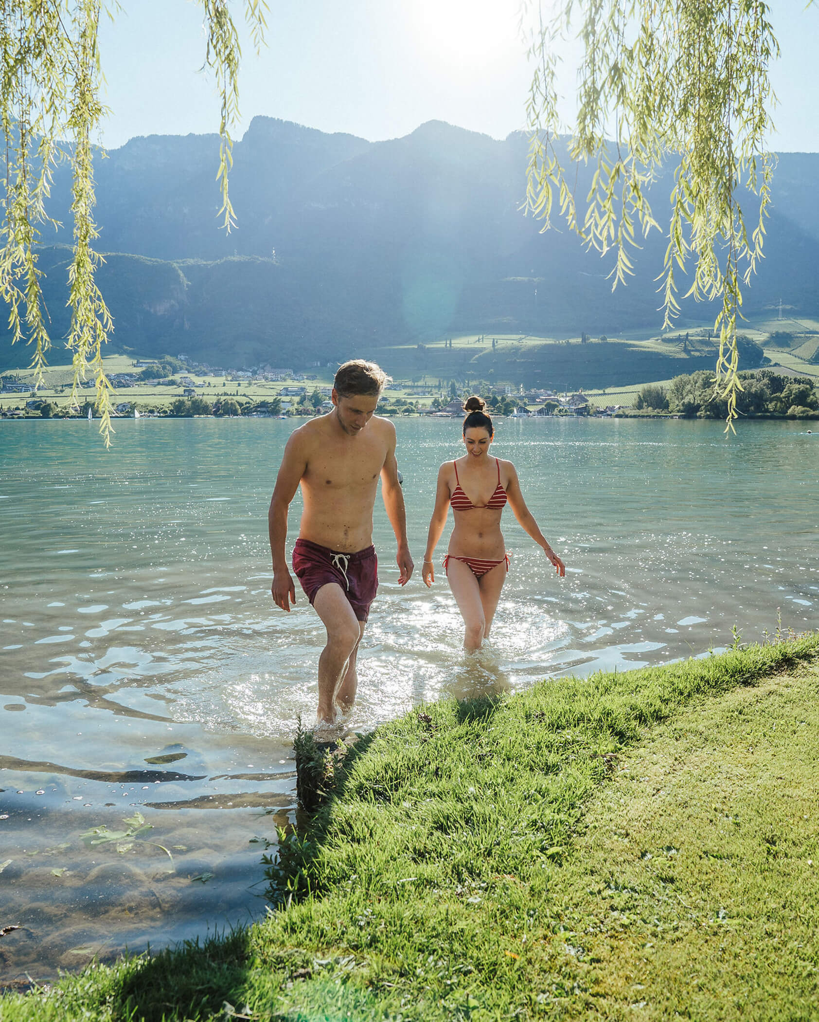 A man and woman in swimsuits walk from a lake onto grassy shore, mountains visible in the background. - Hotel Tenz