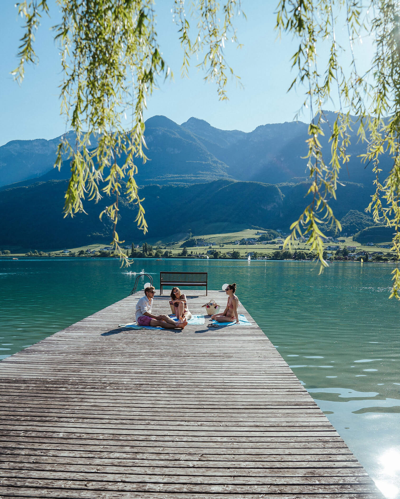 Three people relax on a wooden dock over a clear lake, surrounded by mountains and hanging tree branches. - Hotel Tenz
