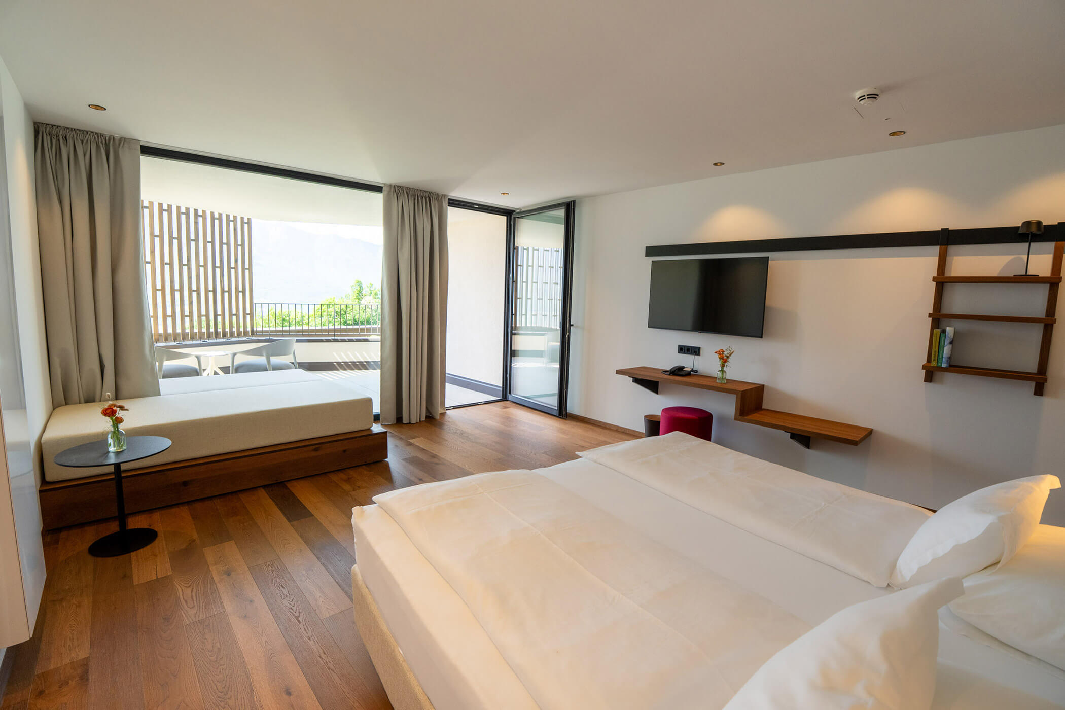 Modern hotel room with large bed, wooden floor, wall-mounted TV, desk, and balcony with outdoor seating. - Hotel Tenz