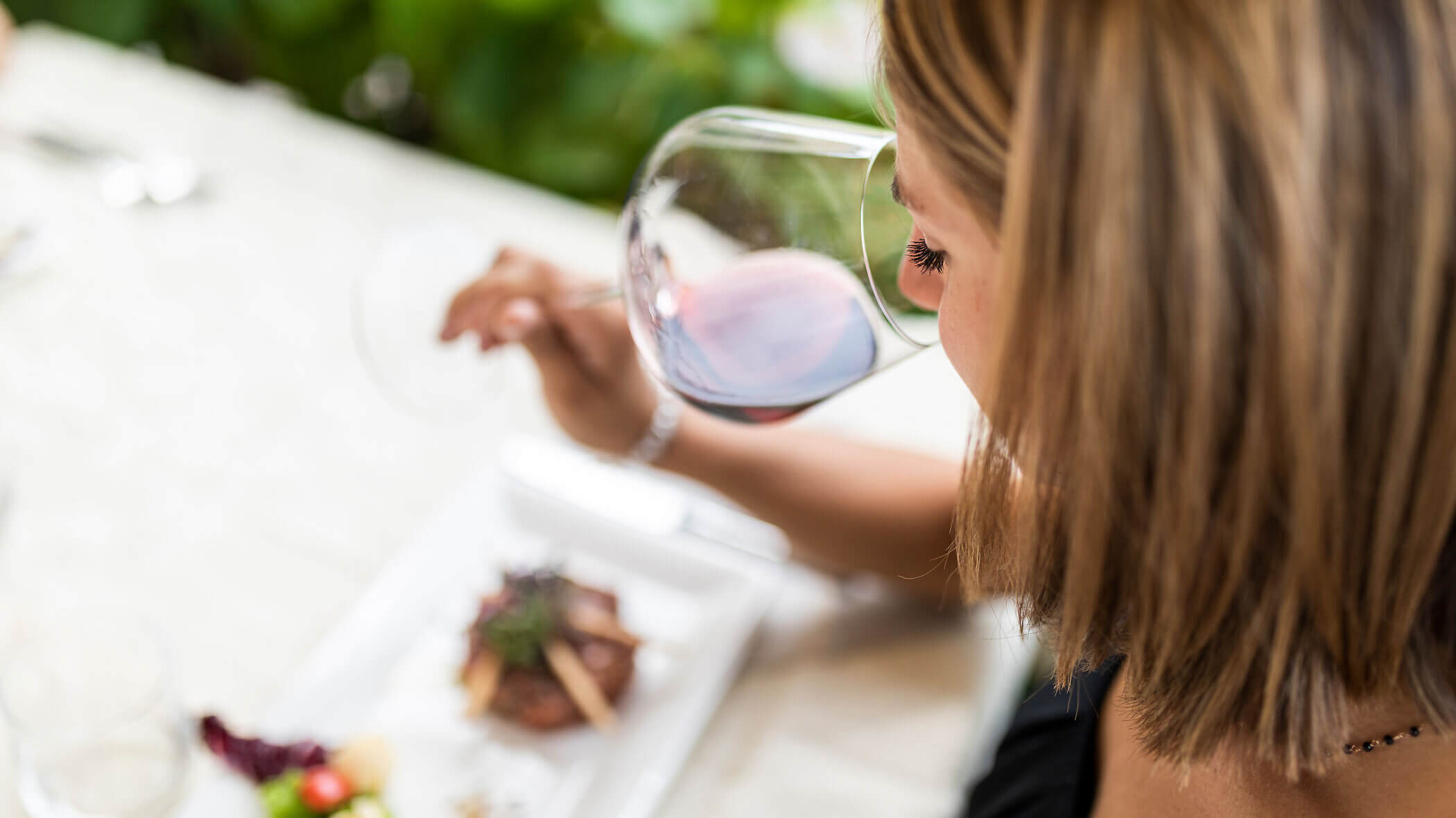A woman with short hair sips red wine at a table set with an elegant meal, viewed from above. - Hotel Tenz