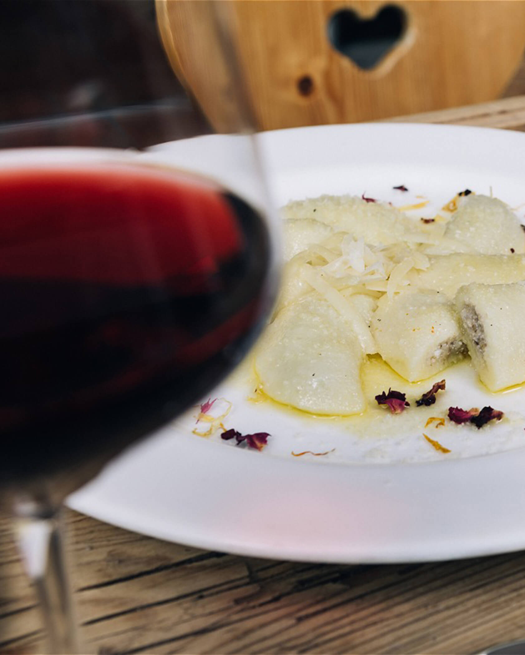 A glass of red wine in front of a plate of ravioli garnished with cheese and flower petals. - Hotel Tenz