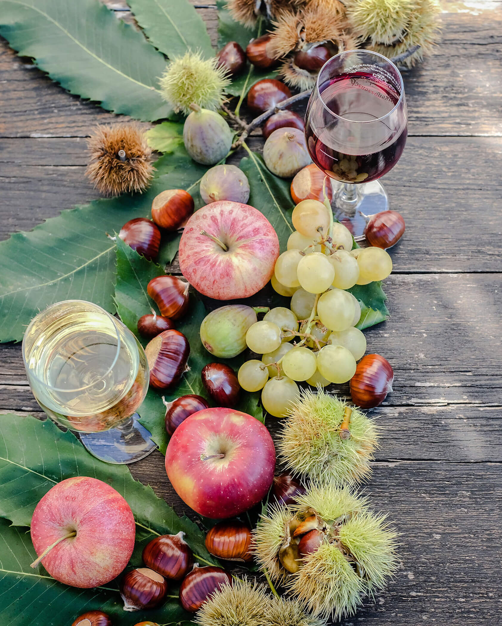 Apples, grapes, figs, chestnuts, and two wine glasses on a rustic wooden table with green leaves. - Hotel Tenz