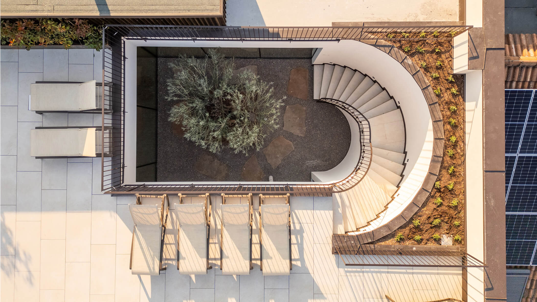 Curved staircase around a tree, with lounge chairs and planters on a tiled patio, viewed from above. - Hotel Tenz