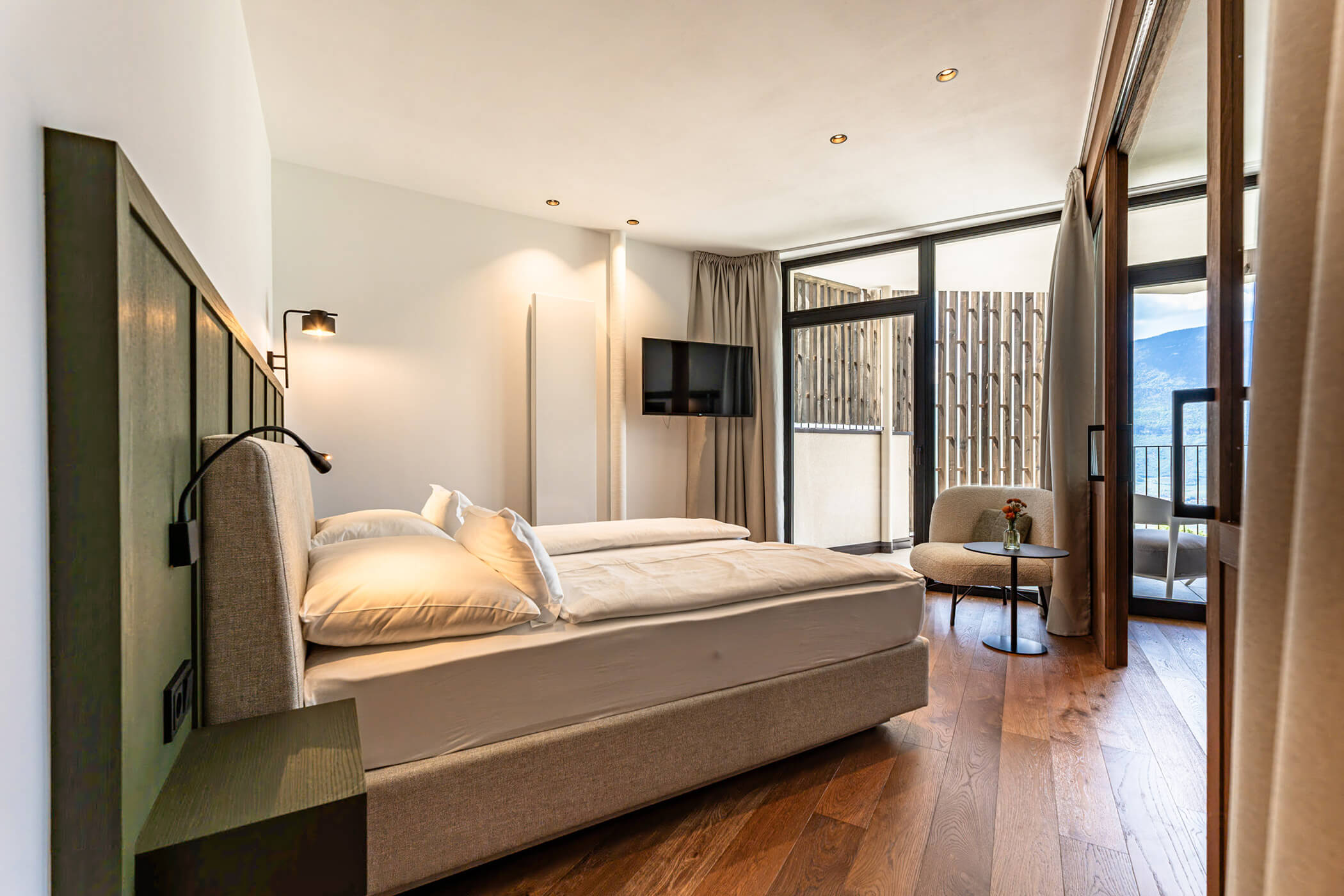 Modern hotel room with two beds, a chair, TV, wood floor, large windows, and a scenic mountain view. - Hotel Tenz