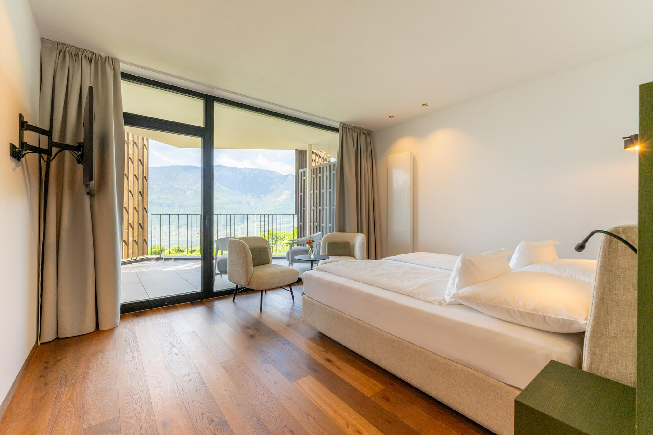Modern hotel room with double bed, chairs, and balcony overlooking mountains and greenery. - Hotel Tenz