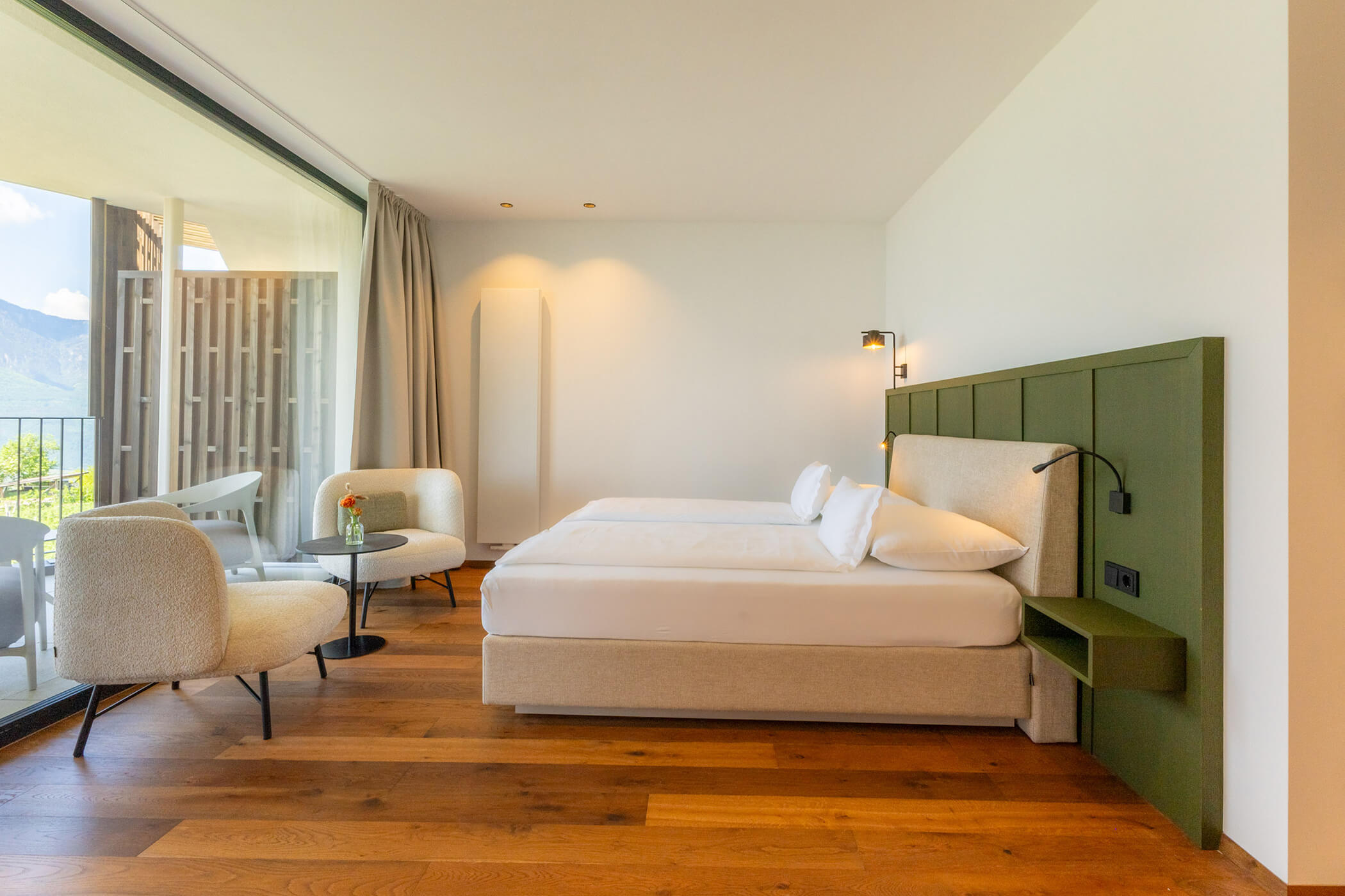 Modern hotel room with a large bed, two armchairs, a small table, and a balcony with mountain views. - Hotel Tenz