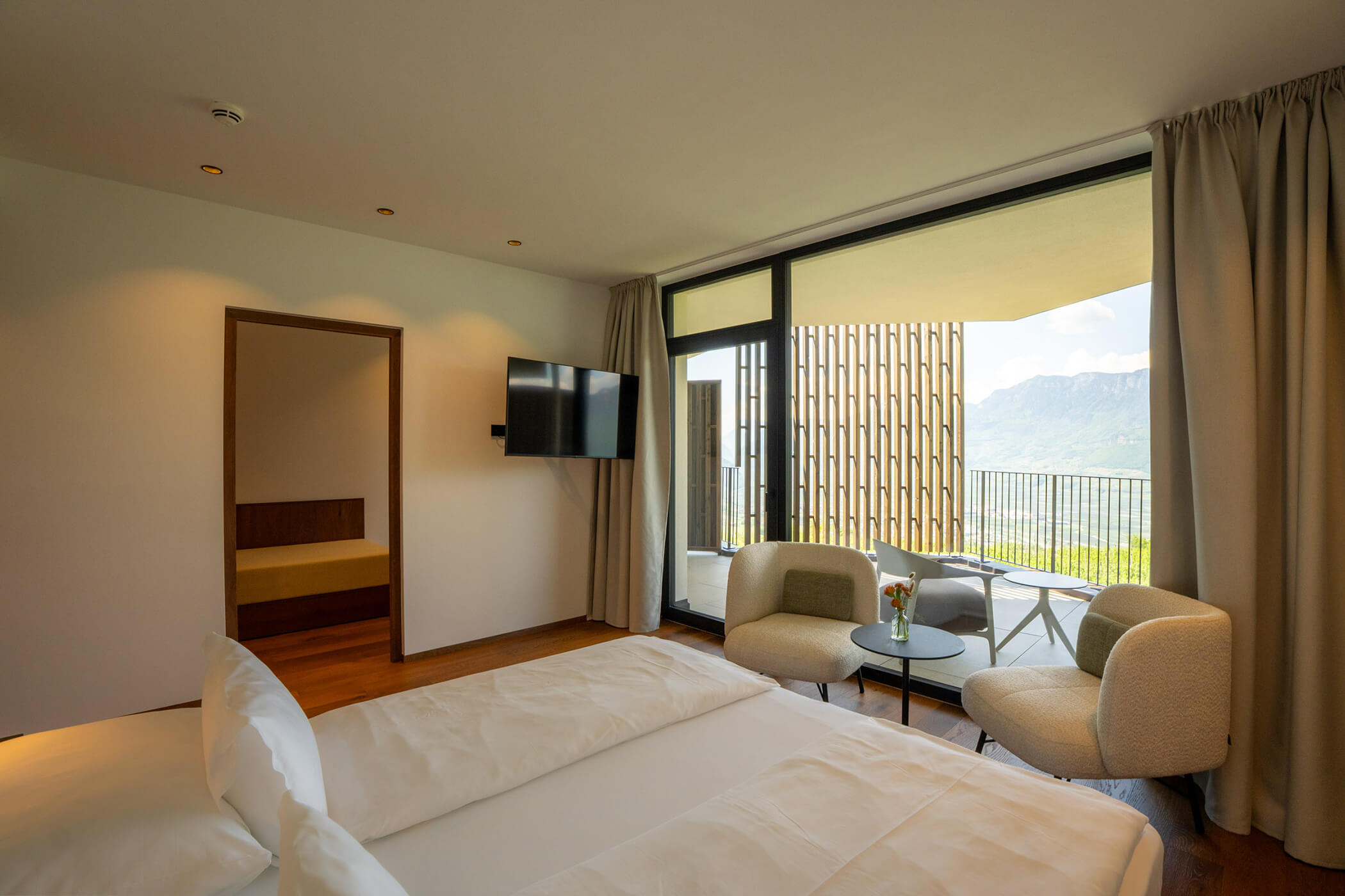 Minimalist hotel room with a bed, seating area, TV, balcony, and mountain view through large windows. - Hotel Tenz