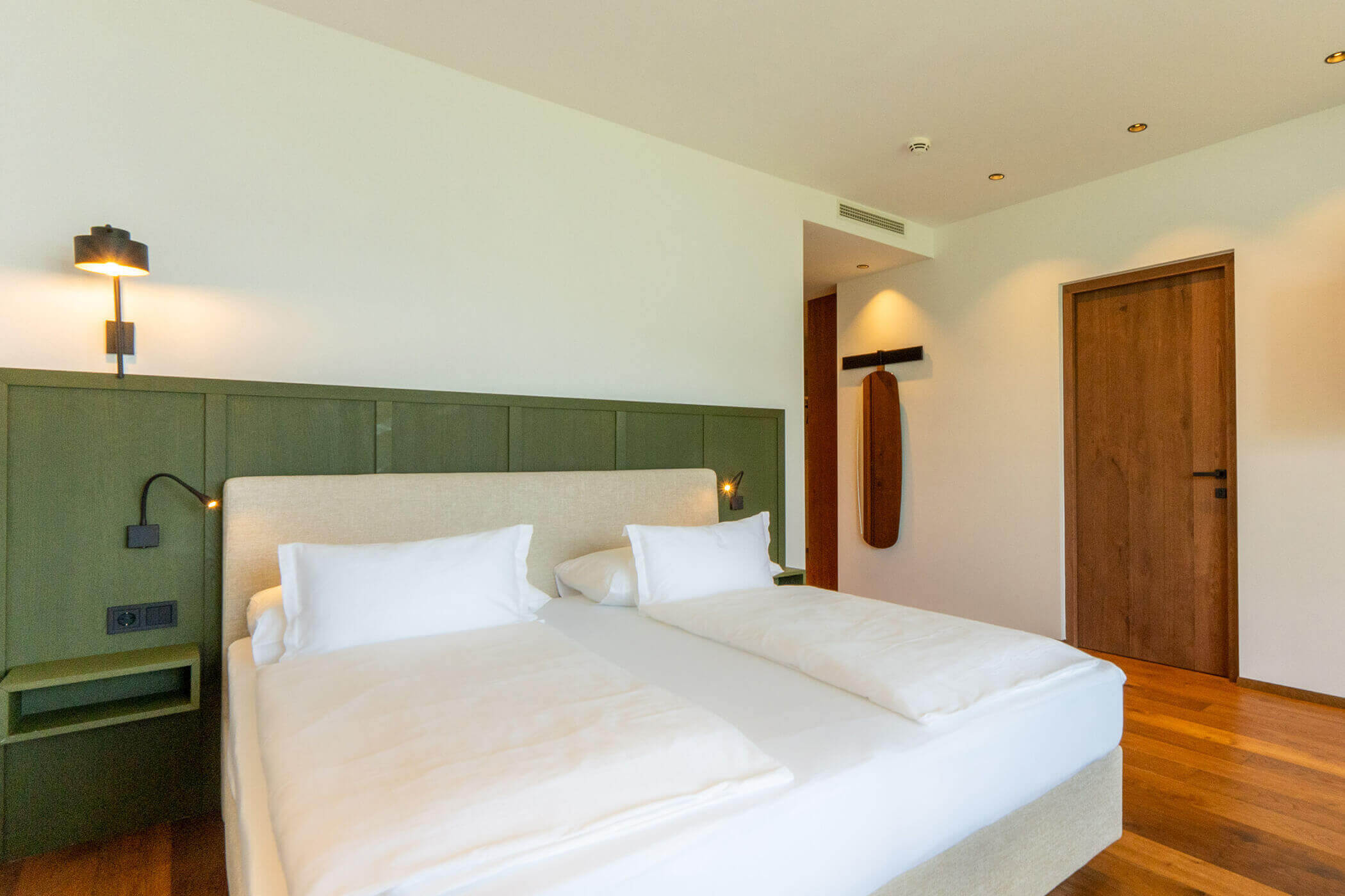 Minimalist hotel room with a double bed, wooden floor, wall lamps, and a closed wooden door. - Hotel Tenz